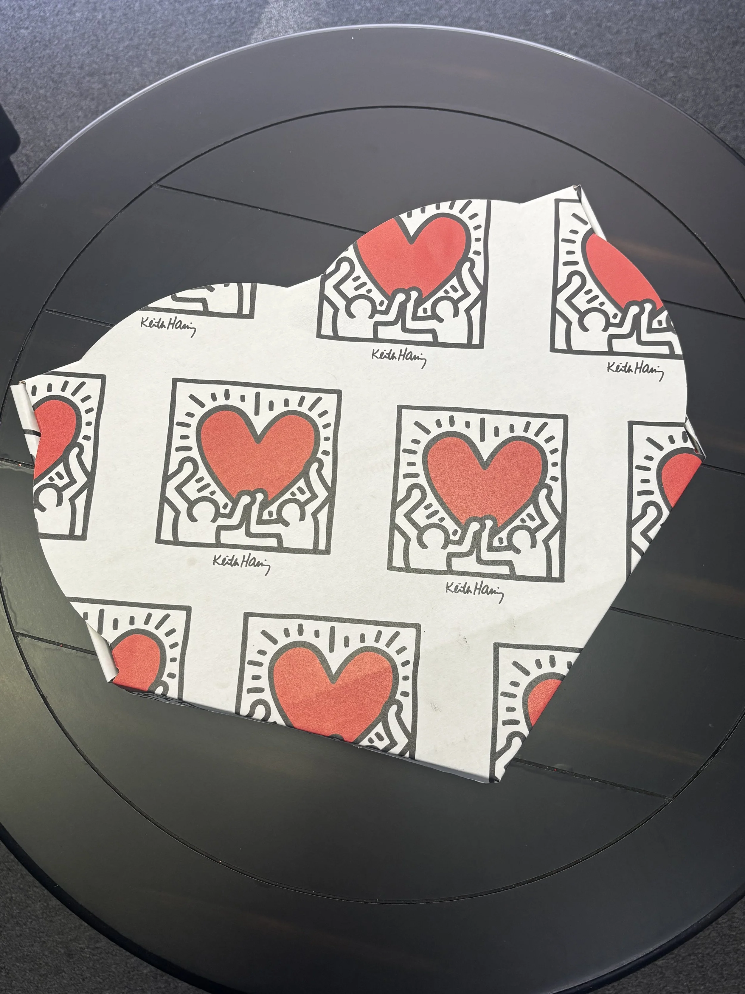 A black circular table with a piece of paper featuring multiple repeating images of stylized hearts and figures, signed 'Keith Haring'.