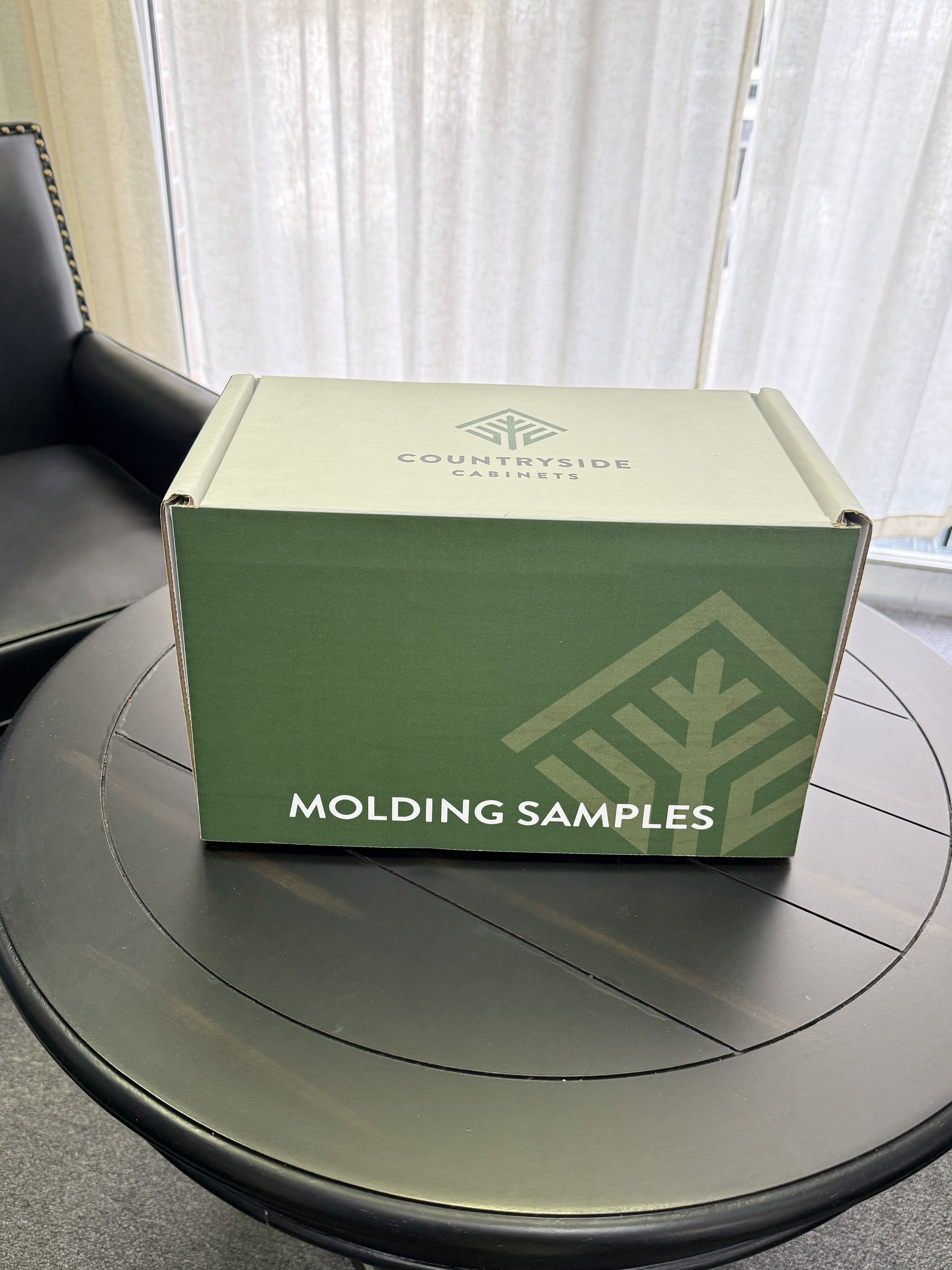 A green cardboard box labeled 'CountrySide Cabinets' with the words 'Molding Samples' is placed on a round black table.