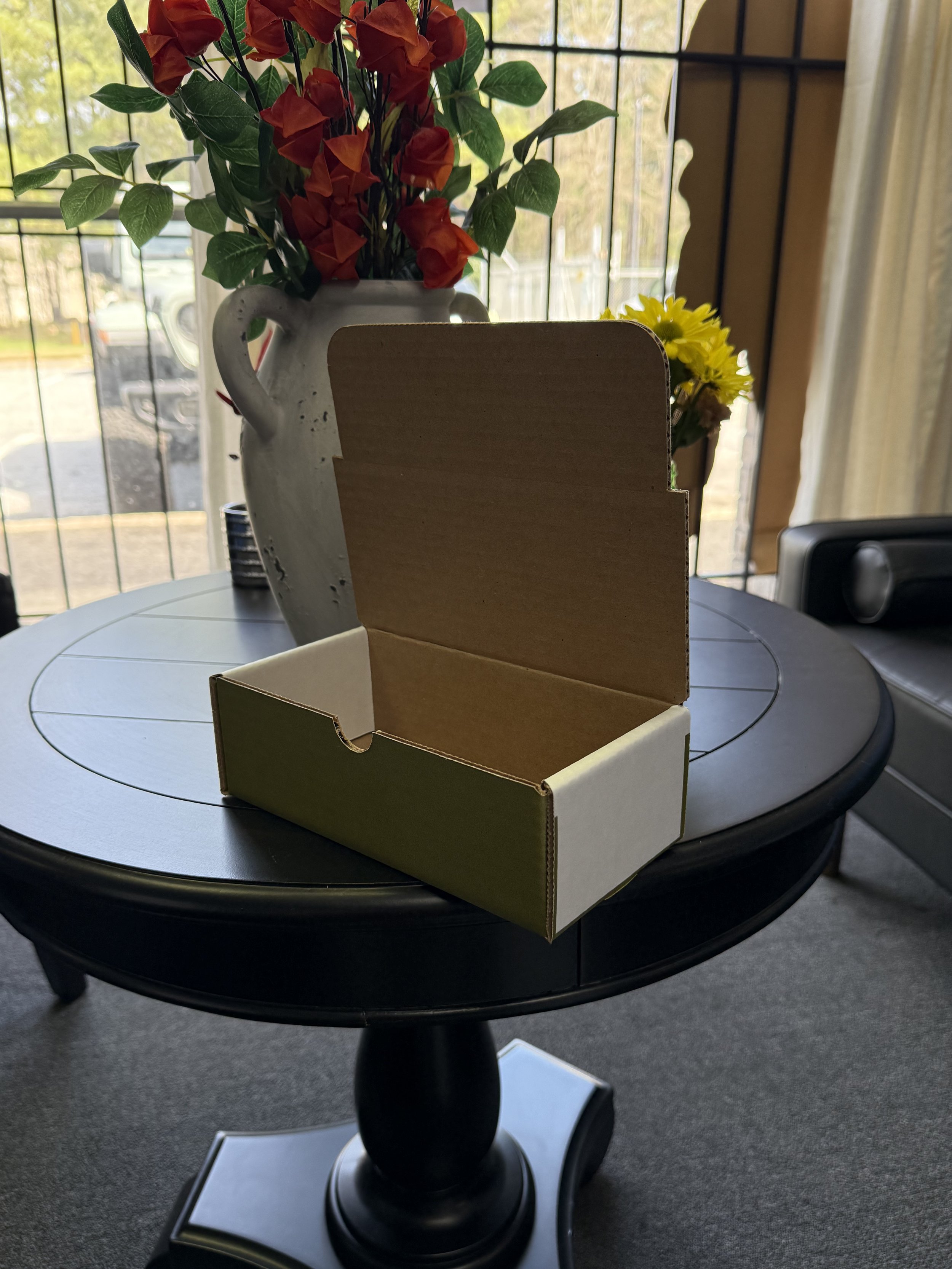 Empty cardboard box on a round black table with a vase of red and orange flowers and yellow flowers in the background, near a window with a view of a parked car and trees outside.
