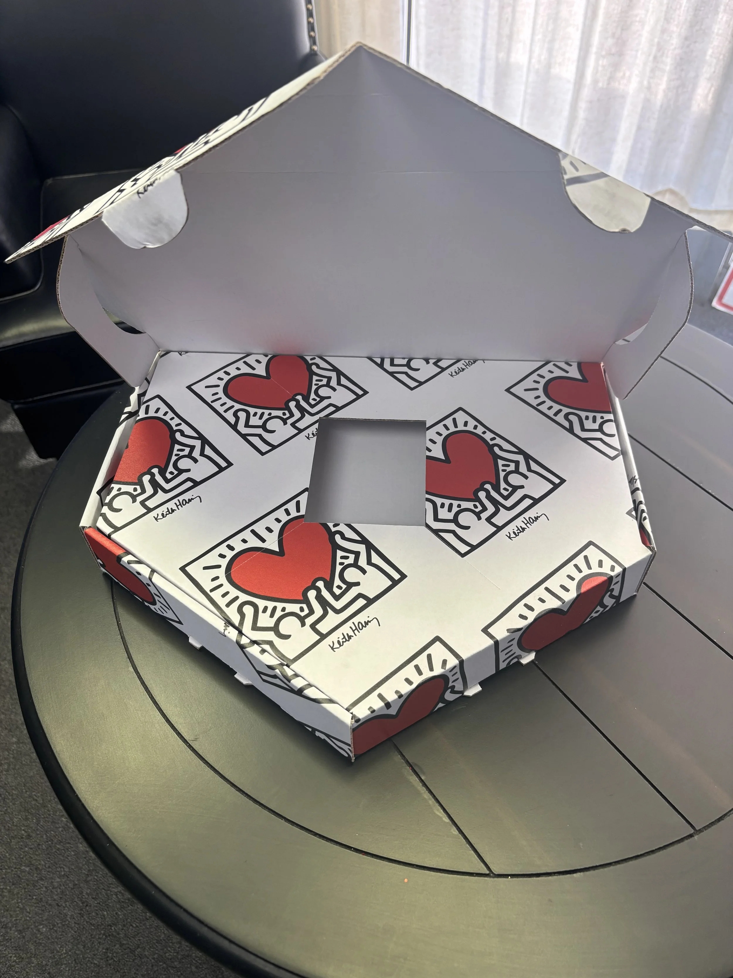 Empty pizza box with a red and black love-themed design, open on a dark green round table near a window.