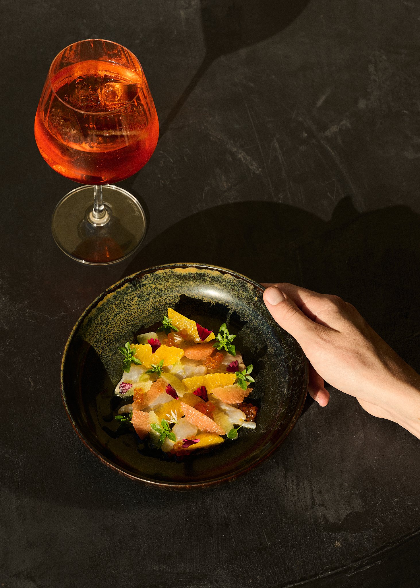Une assiette avec un plat à base de poisson, garnie de petites fleurs et d'herbes, accompagnée d'un verre de boisson rosée avec glaçons sur une table sombre.