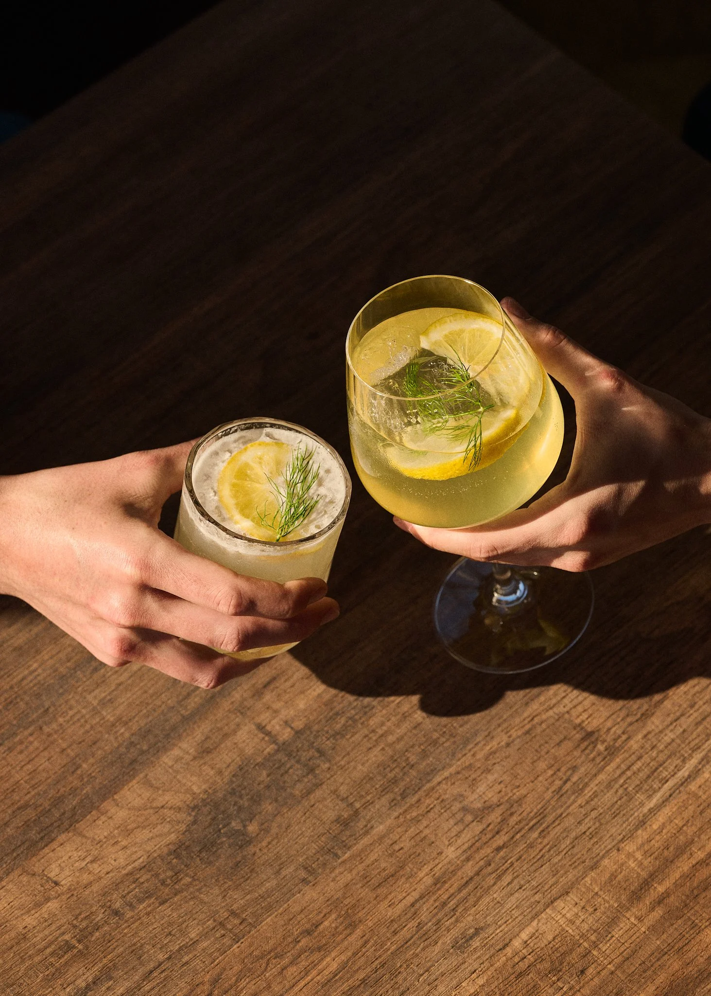 Deux mains tiennent des verres à cocktail contenant des boissons citronnées avec des tranches de citron, des glaçons et des herbes fraîches, sur une table en bois.