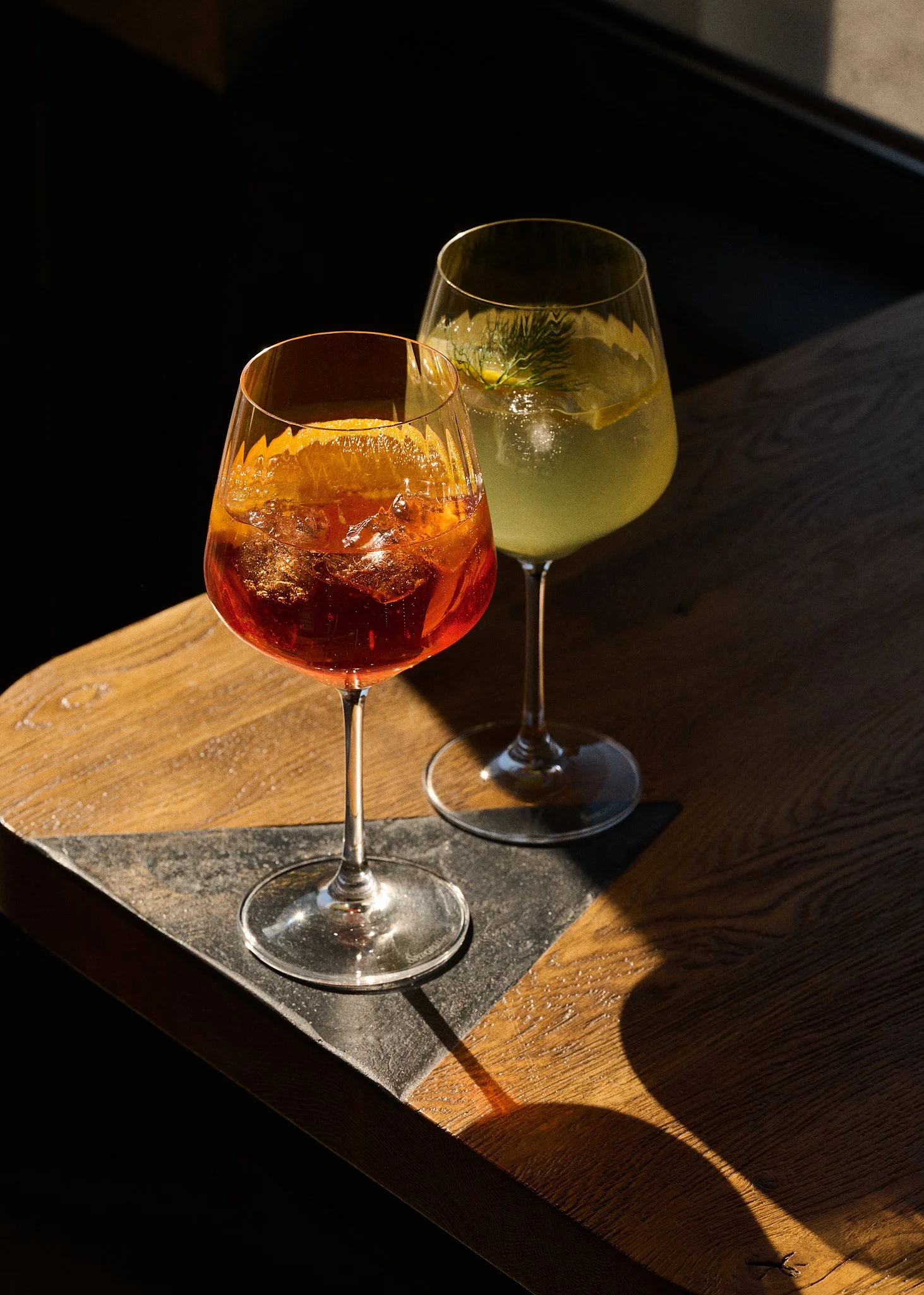 Deux verres à cocktail, l'un rouge avec des glaçons, l'autre vert avec une branche de décor, placés sur une surface en bois avec un fond sombre.