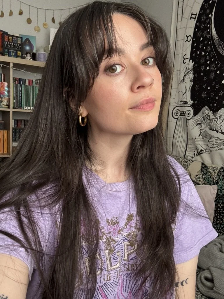 A woman with long dark hair and hoop earrings, wearing a purple graphic T-shirt, taking a selfie in a room with bookshelf and tapestry in the background.