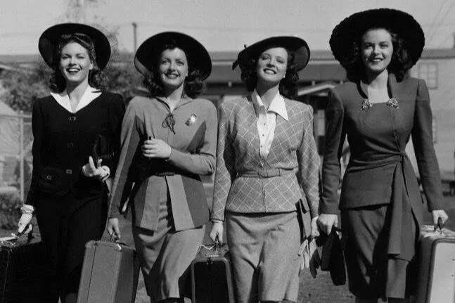 Cuatro mujeres vestidas con ropa de estilo vintage, llevando sombreros y maletas, caminando juntas al aire libre.
