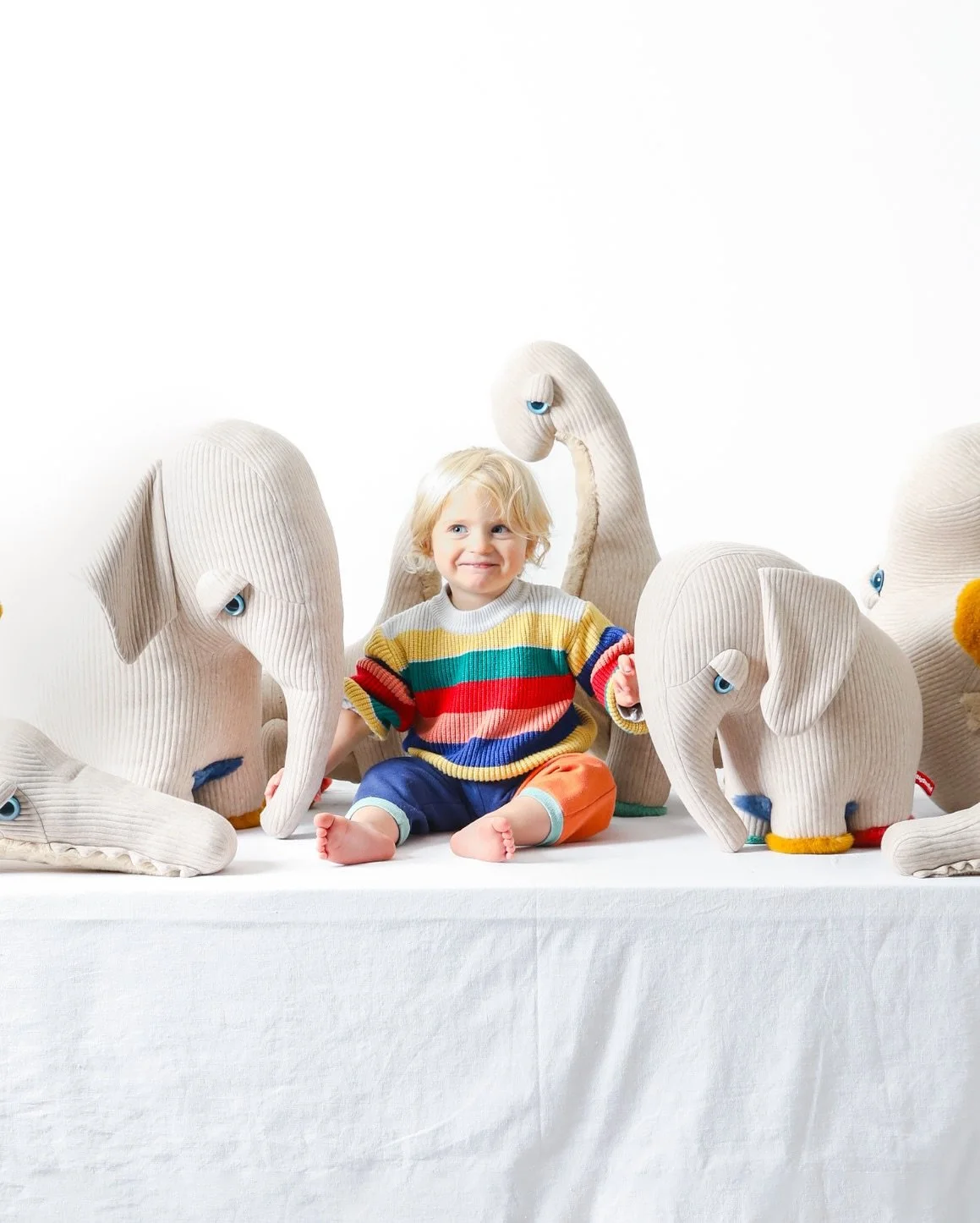 Niño sonriente sentado entre peluches de elefantes y jirafas de tela con expresiones amigables, en un fondo blanco y superficie blanca.