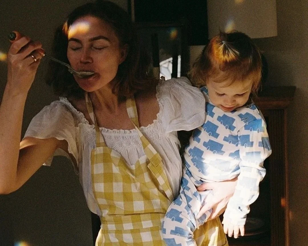 Mujer con delantal amarillo y vestido blanco disfrutando comida con un niño pequeño con pijama azul y blanca en un interior