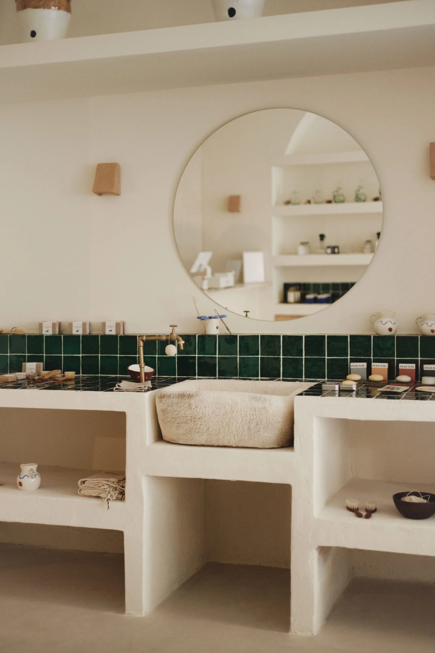 Una vista de una pared interior con un espejo circular y estanterías decorativas blancas que contienen pequeños objetos y vajilla, con un fregadero y accesorios de baño en primer plano.