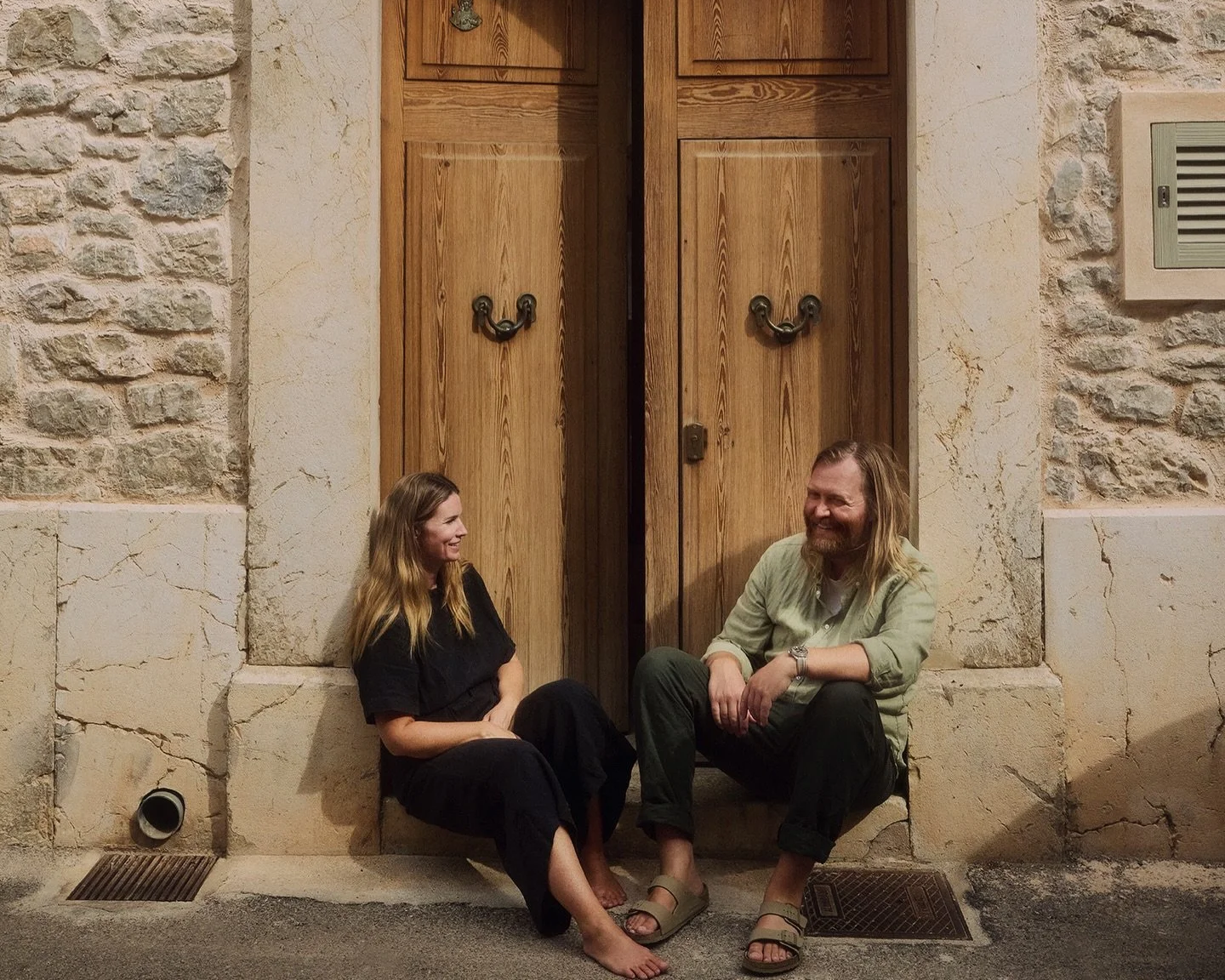 Dos personas sentadas en el suelo frente a una puerta de madera en una pared de piedra, conversando y sonriendo.