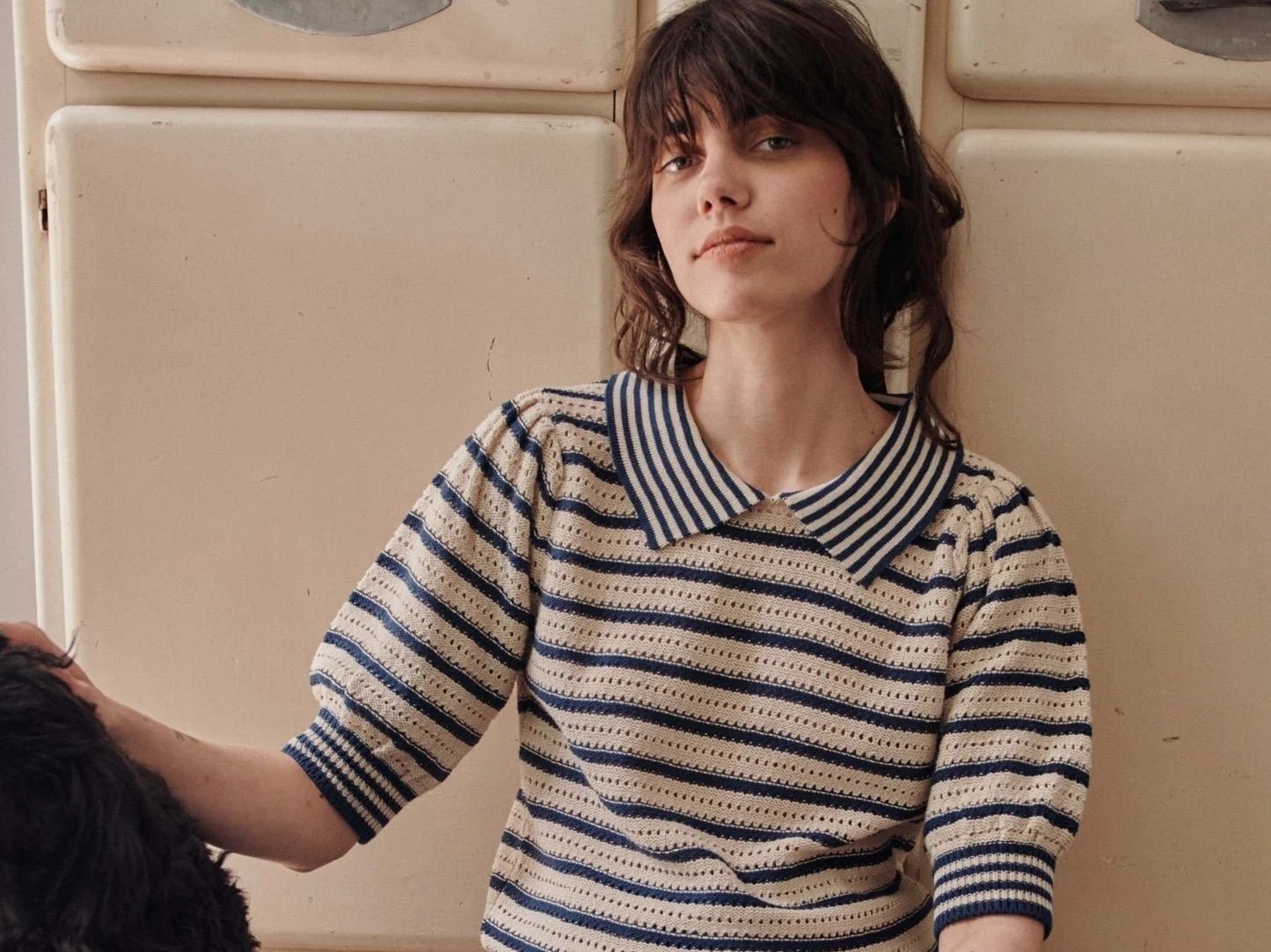 Retrato de una mujer joven con cabello castaño oscuro, vestida con un suéter a rayas y un cuello de camiseta a rayas, sonriendo suavemente, apoyada contra un fondo de paneles beige.