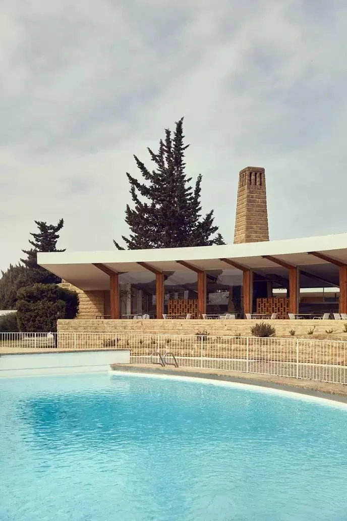 Piscina en primer plano con estructura moderna y chimenea de ladrillo en el fondo, rodeada de árboles y césped.