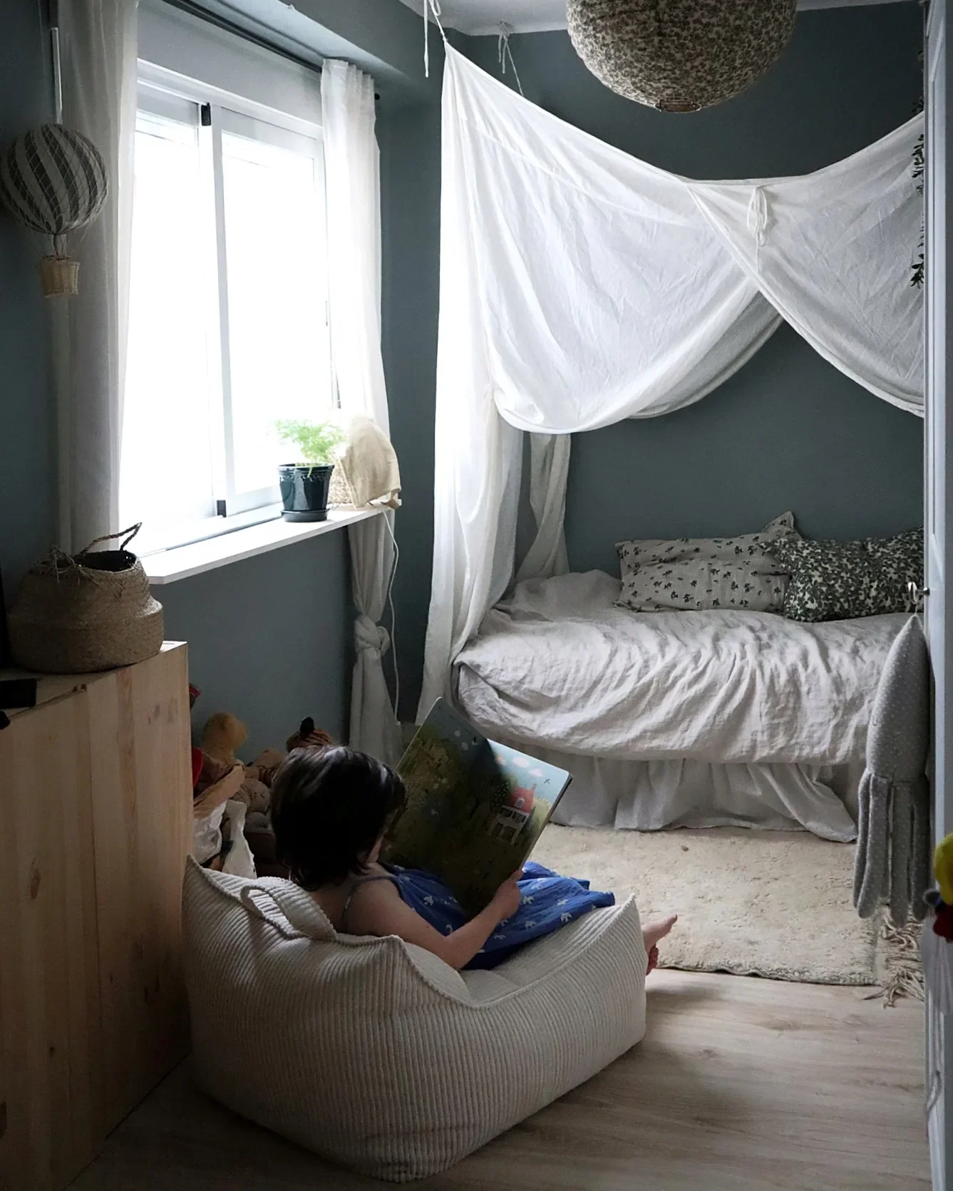 Una habitación con cama con mosquitero, ventana con cortinas blancas, niños sentados en un sillón y en el suelo, uno leyendo un libro, adornos y plantas en la habitación.