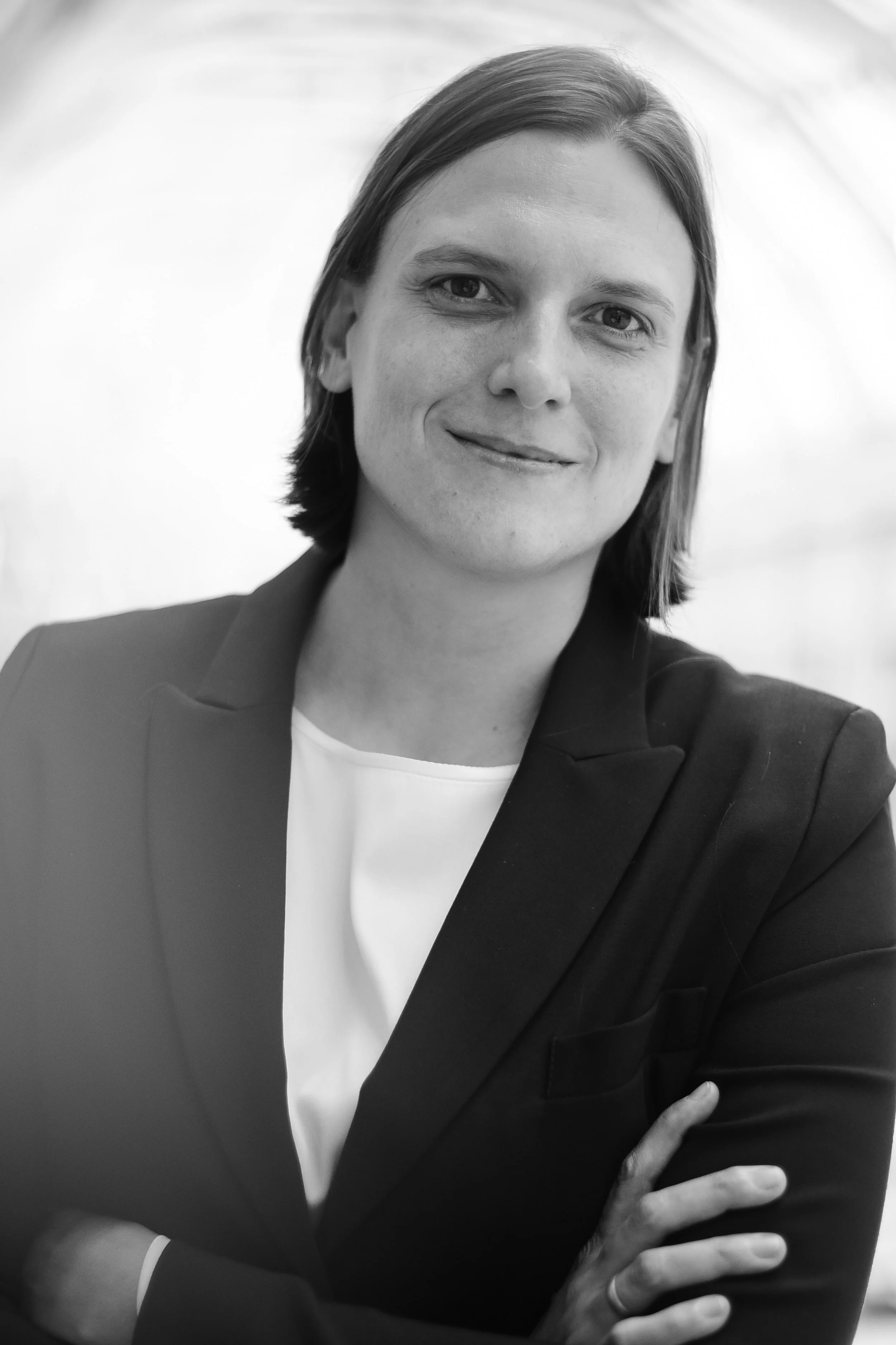 Schwarz‑weißes Oberkörperporträt einer Person mit leicht verschränkten Händen vor dem Körper, neutraler Hintergrund.