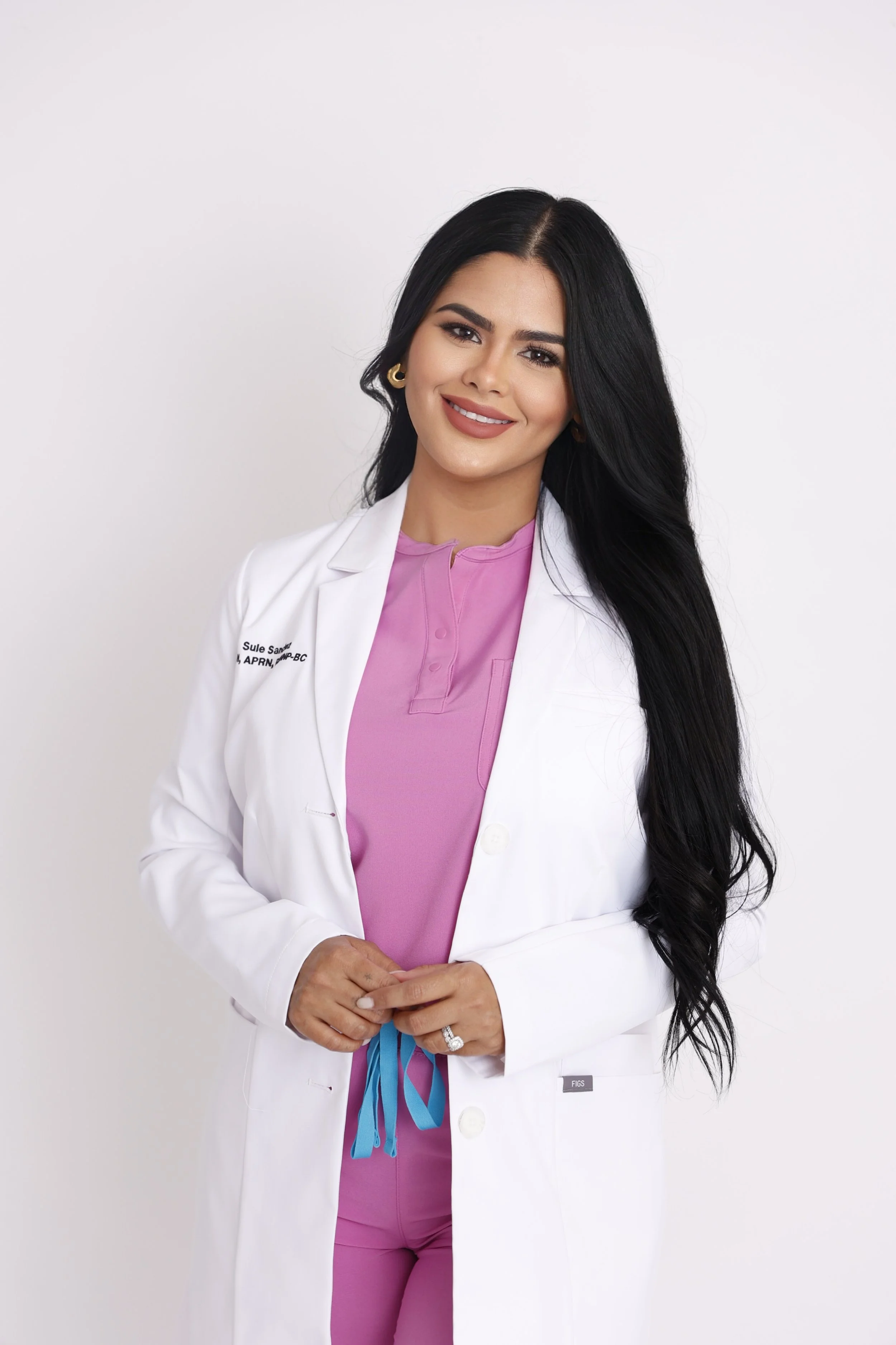 A woman with long black hair wearing a white coat over pink scrubs, standing against a white background.