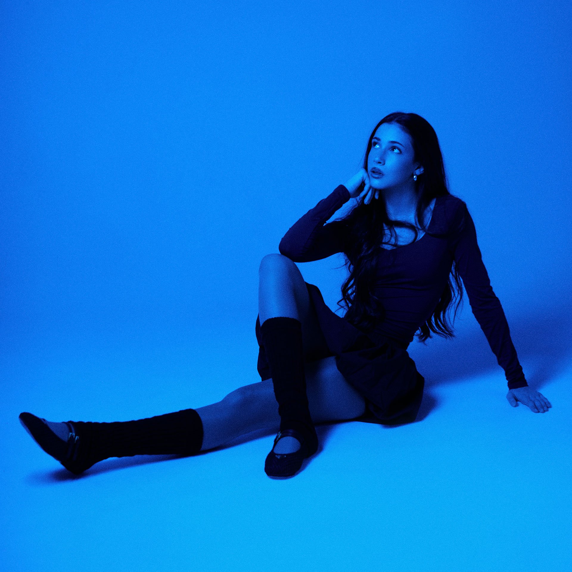 A young woman with long dark hair and earrings, dressed in a dark long-sleeve top and skirt, sitting on the floor with one knee bent and leg extended, wearing knee-high socks and flat shoes, against a blue background, looking thoughtfully upwards to her left.
