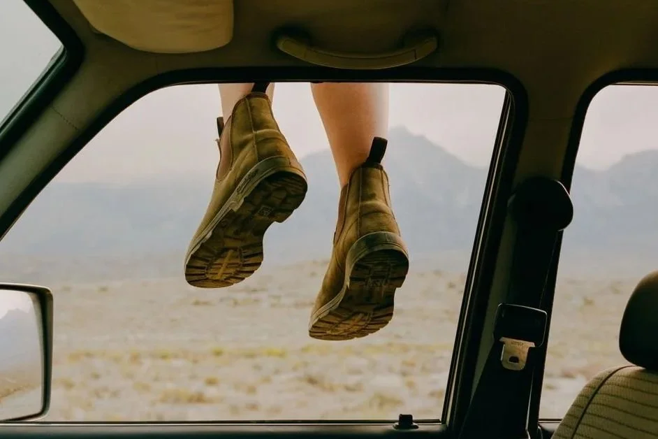 Scarpe da trekking appoggiate fuori da un finestrino di un'auto in un paesaggio desertico con montagne in lontananza.