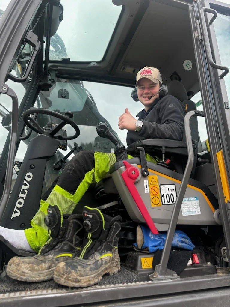 Ein ung mann i arbeidskle som sit i ei gravemaskin og smiler, med tommel opp.