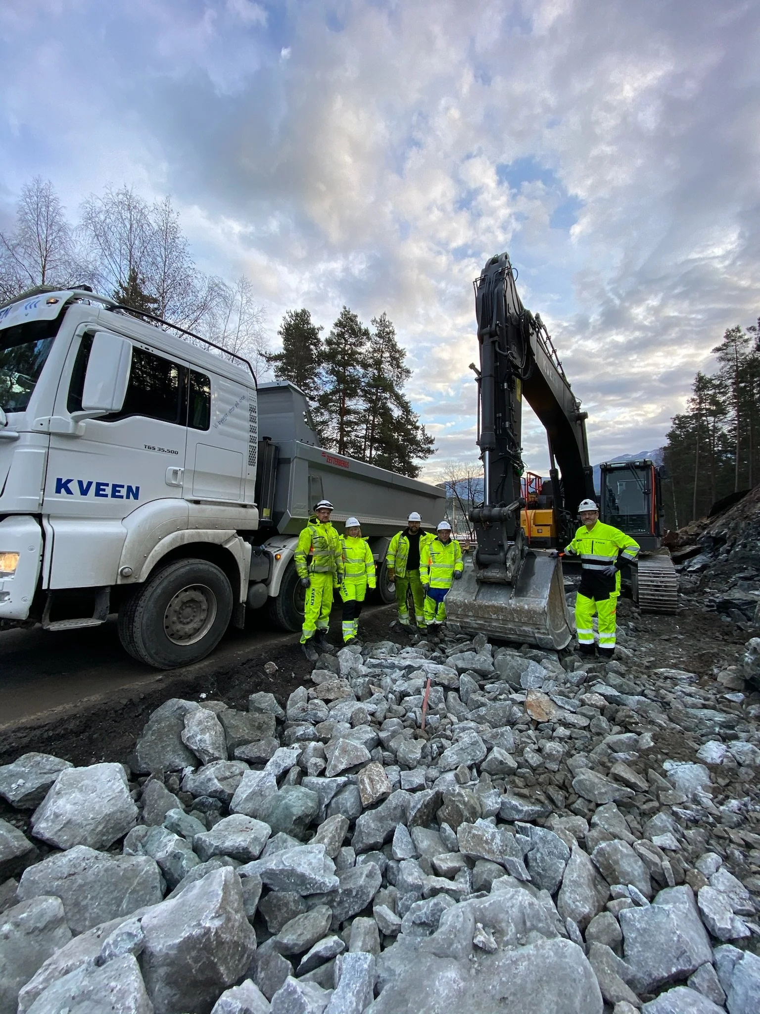 Anleggsarbeidarar i arbeidskle og hjelm står ved sidan av ein stor kvit lastebil og ei gravemaskin på eit anleggsområde.