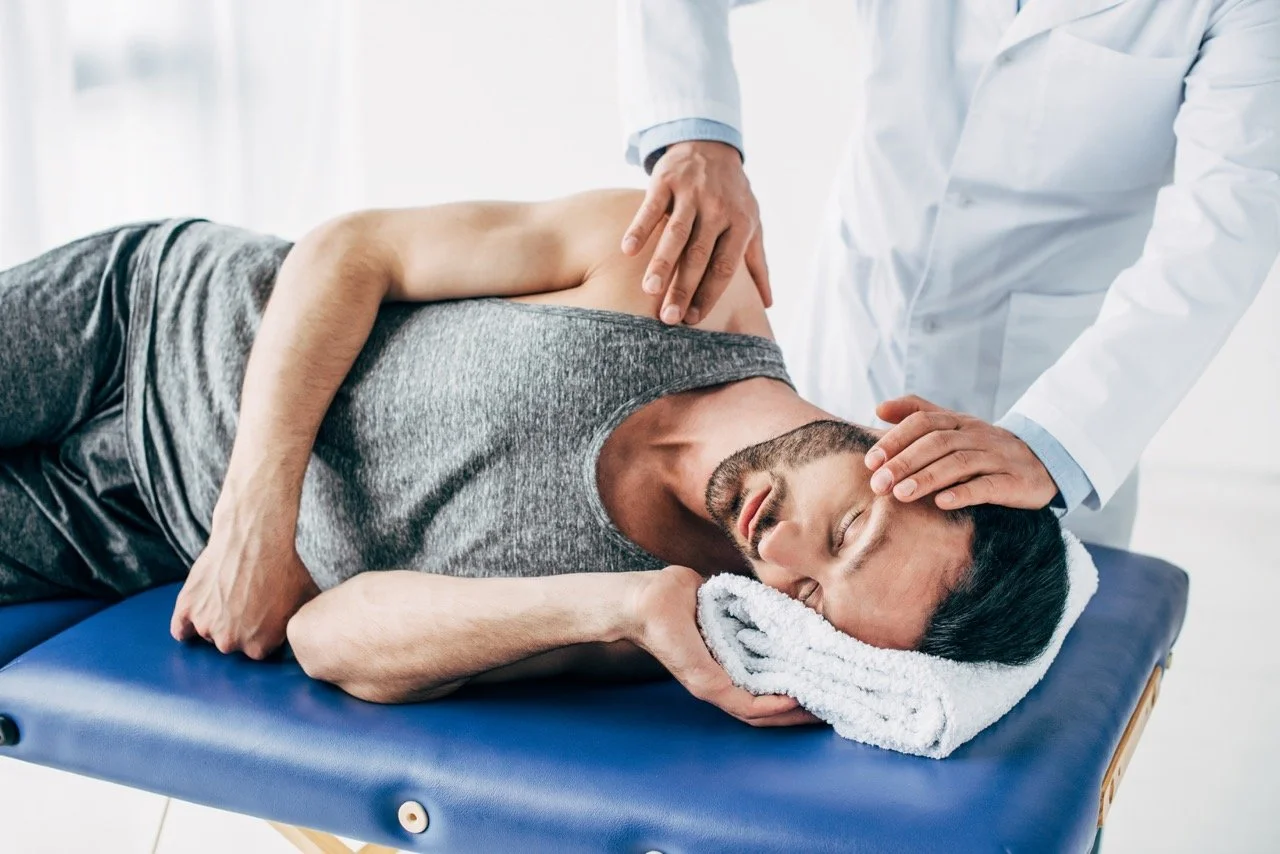 A man lying face down on a medical examination table, receiving a chiropractic or physical therapy treatment from a healthcare professional in a white coat.