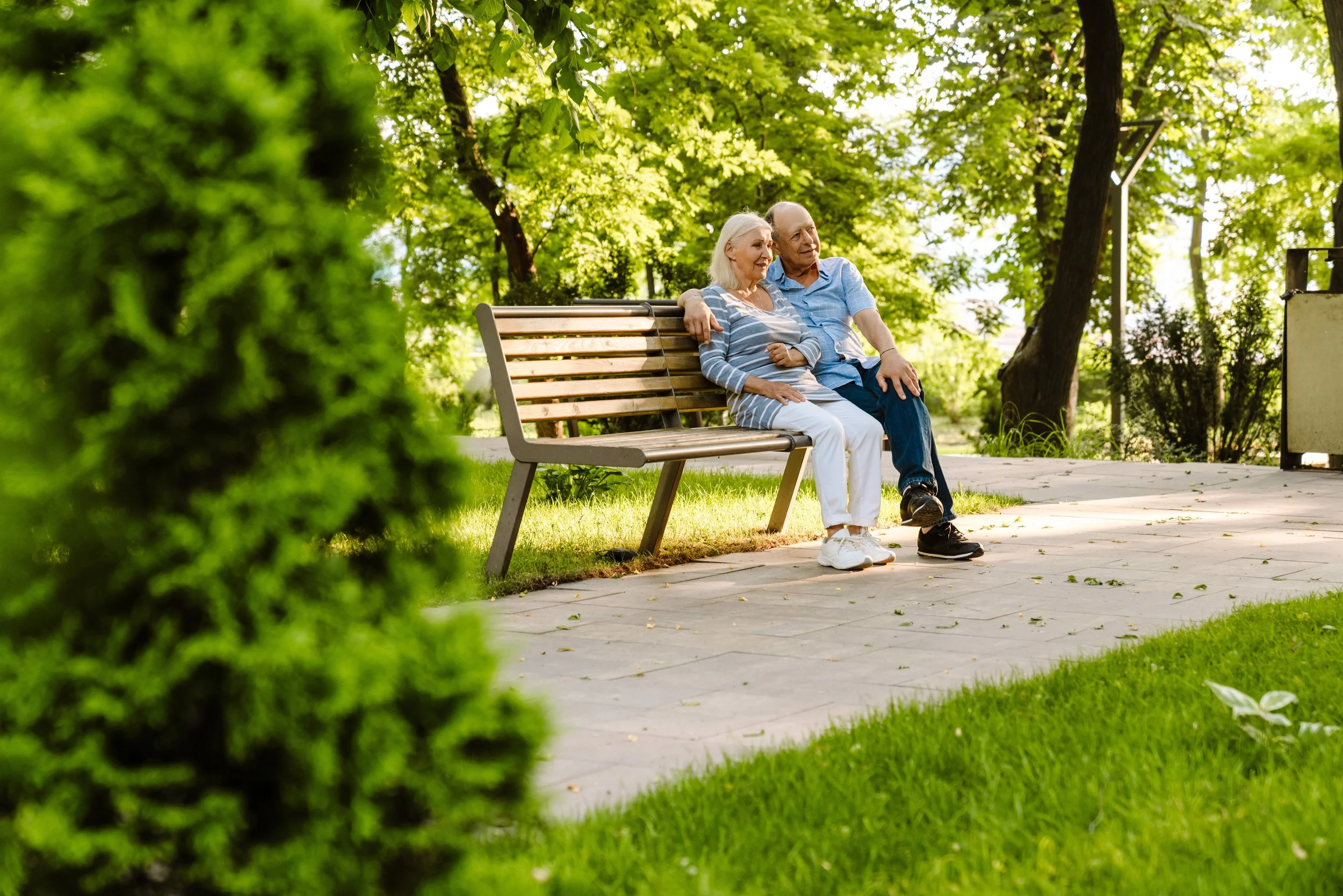 An elderly couple sitting closely together on a park bench, surrounded by green trees, enjoying a peaceful moment.
