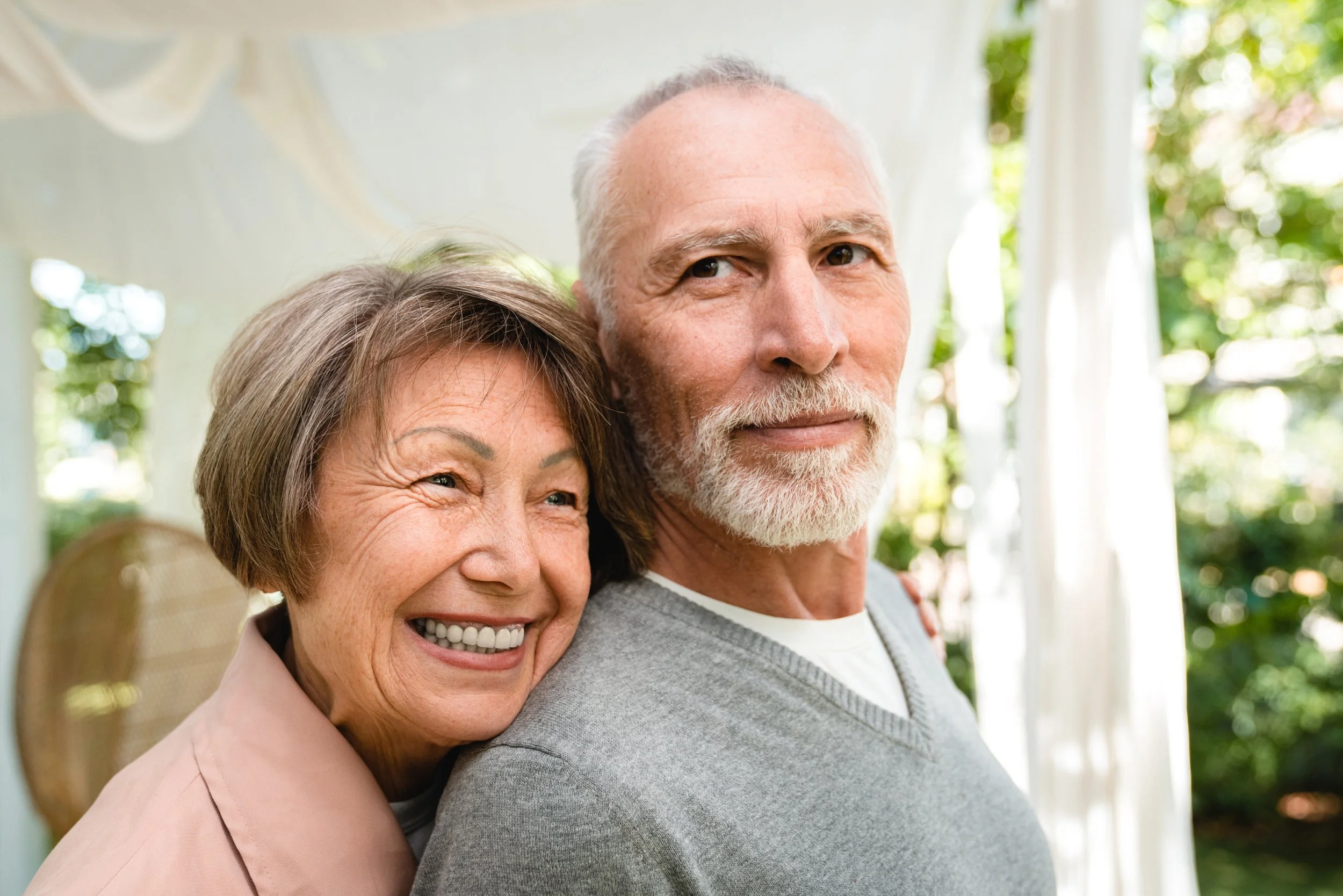 Older woman and man smiling outdoors in a garden, with the woman hugging the man from the side.