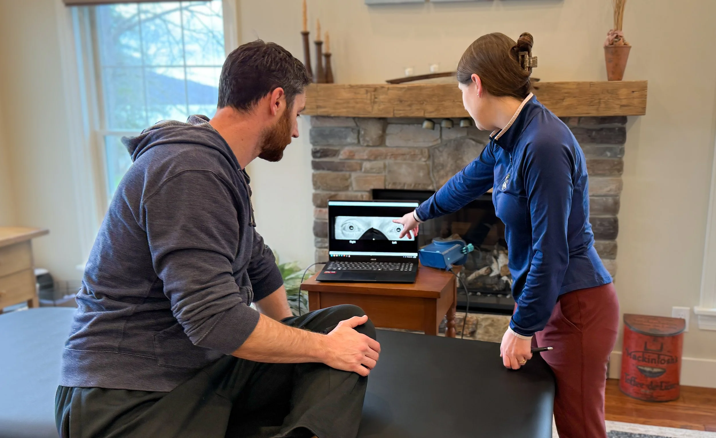 A healthcare professional projecting eye test results on a screen using a handheld instrument for a male patient sitting on an examination table in a cozy room with a fireplace, wooden furniture, and a window.