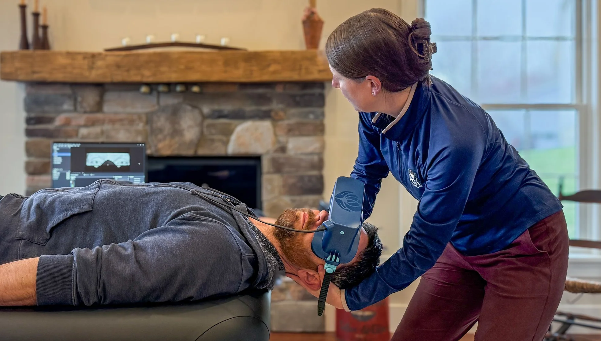 A woman performs CPR on a man lying on a table while using virtual reality goggles, with a computer monitor displaying data in the background.