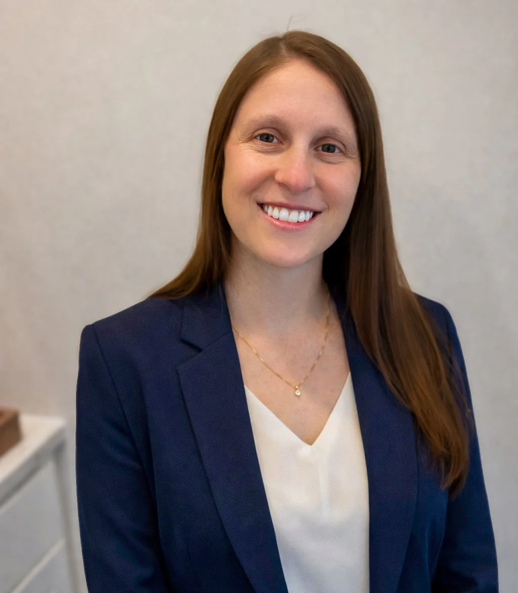 A woman with long brown hair smiling, wearing a navy blazer over a white top and a gold necklace, standing against a plain light-colored background.
