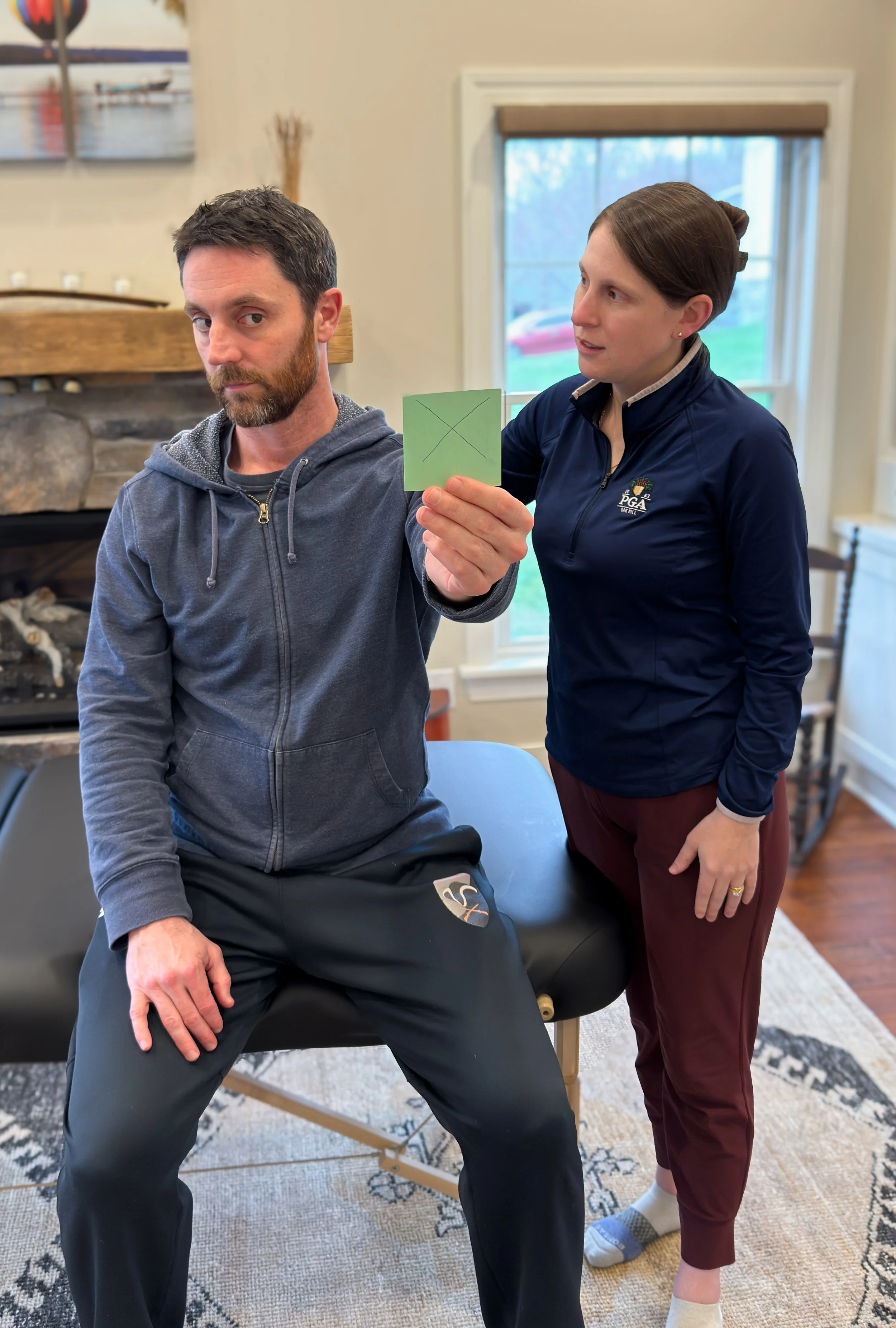 A man sitting on a therapy table with his hand on his knee, holding a green post-it note with a black 'X' on it, while a woman stands next to him, looking at the note. The woman wears a navy jacket and maroon pants, and the man wears a gray hoodie and black pants. The setting is a cozy room with windows, a fireplace, and framed pictures.