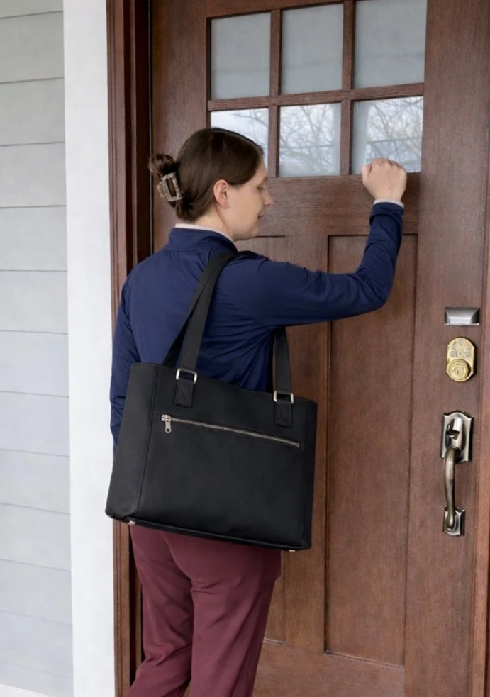 A woman standing outside a door, knocking with her right fist. She has brown hair tied in a bun, and is carrying a black shoulder bag.