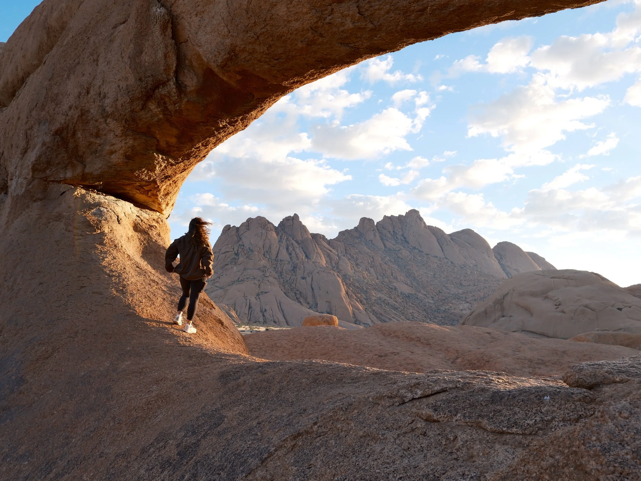 A person hiking in a rocky desert landscape with large rock formations and a canyon, under a partly cloudy sky.