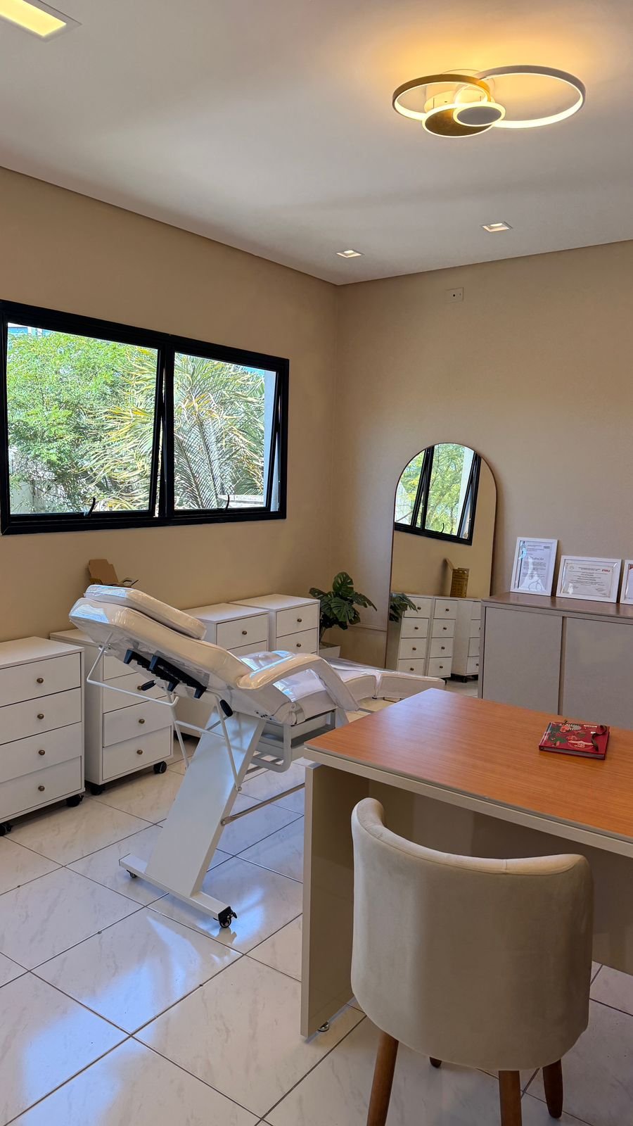 Sala de consulta médica com cama de exame, armários brancos, espelho de arco e planta decorativa, iluminação moderna e janelas com vista para árvores.