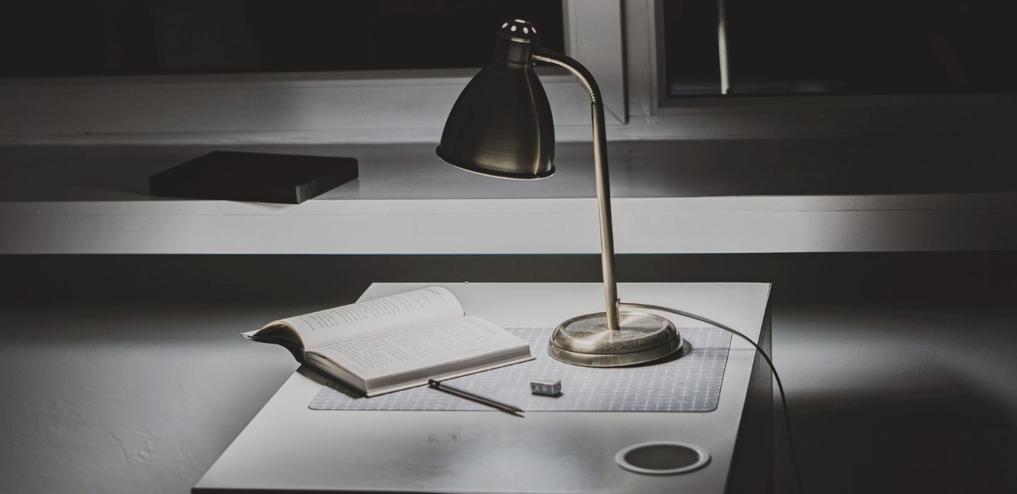 A desk with an open book, a pen, a small eraser, a desk lamp, and a circular cable grommet, with a blurred background.