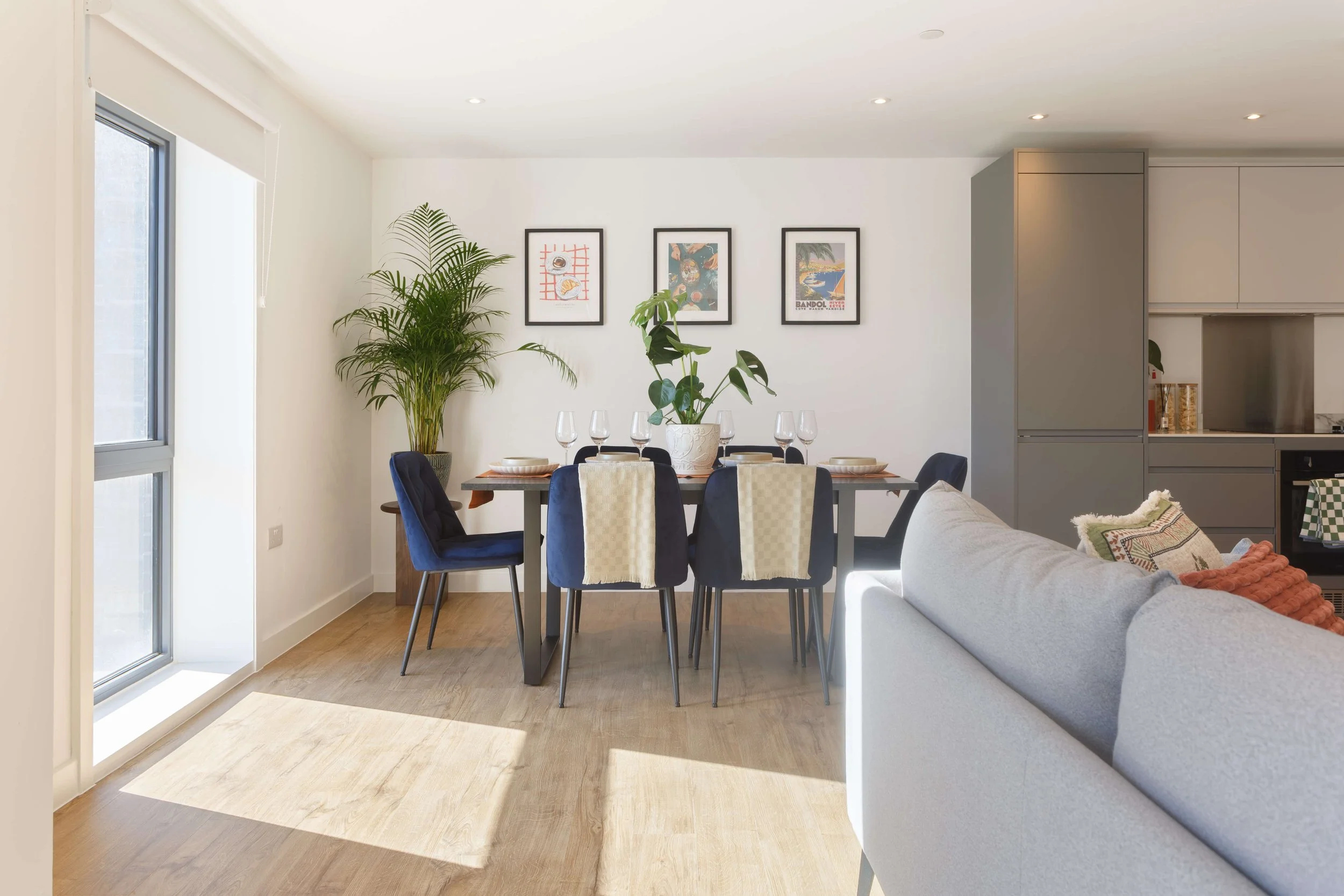 A modern dining area with a white wall, black framed pictures, and plants. A dining table with six chairs and table settings is visible, near a window with natural light.