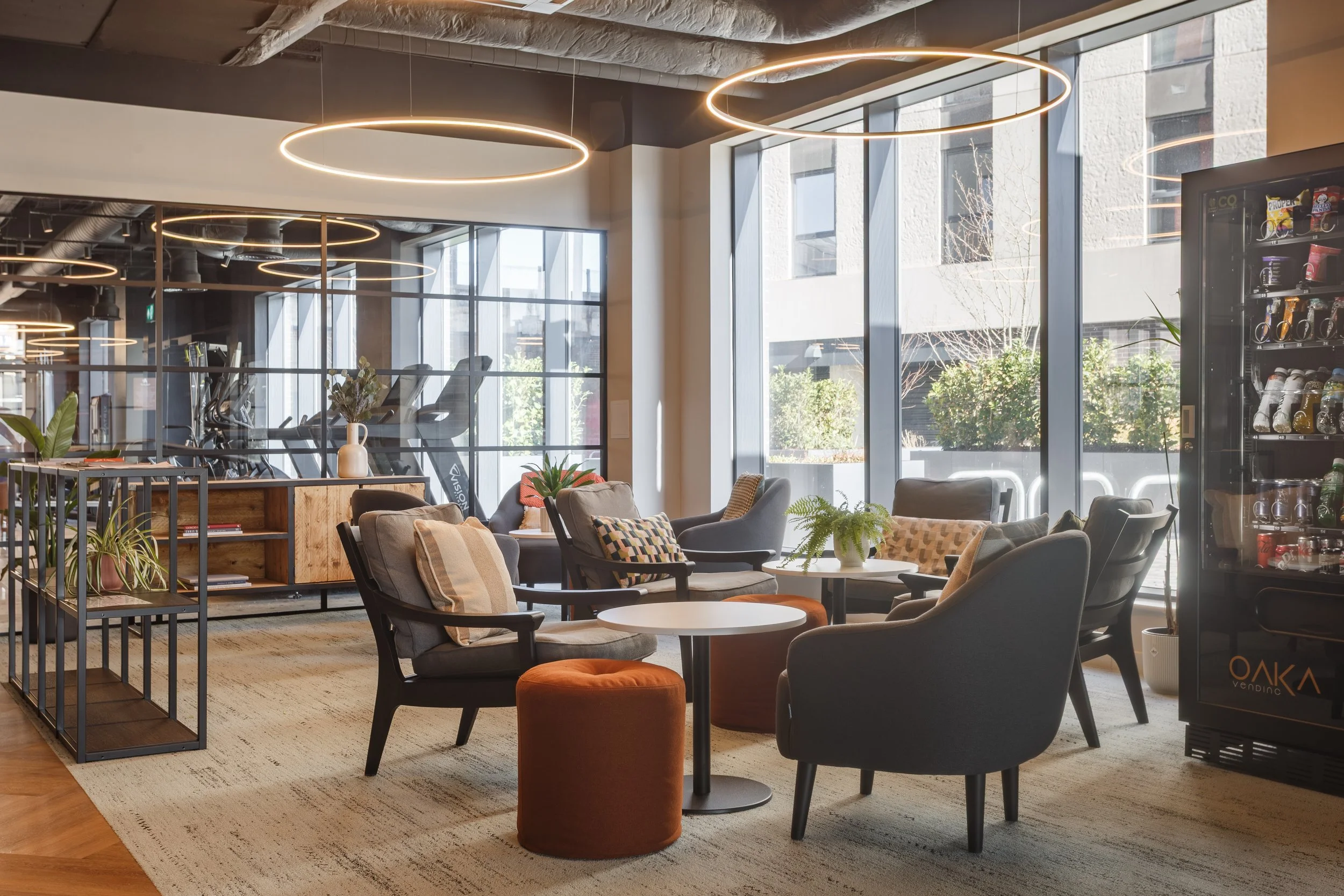 Modern lounge area with chairs, tables, indoor plants, sunlight, circular ceiling lights, large windows, vending machines, and a glass partition.