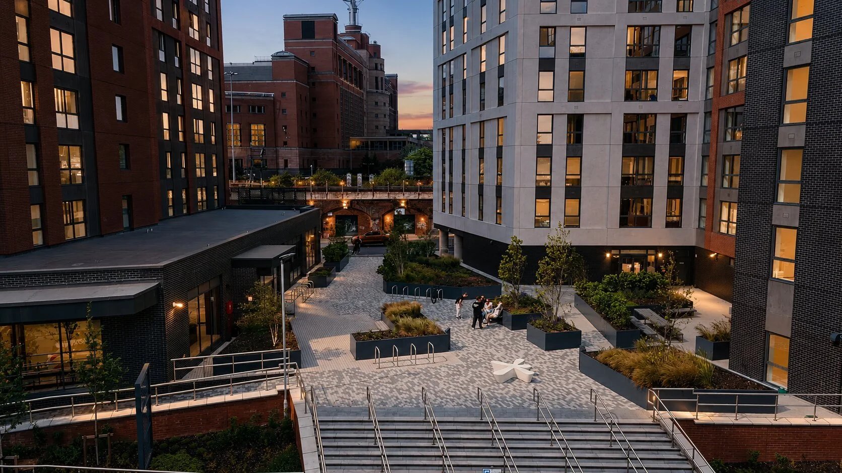 Core of urban apartment complex with landscaped courtyard, seating, and people walking, surrounded by tall modern buildings during twilight.