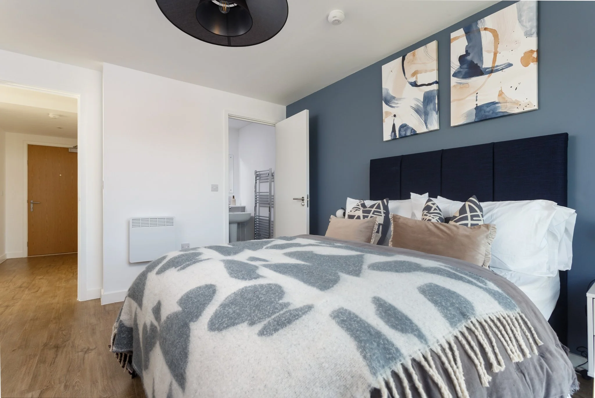 A bedroom with a bed covered in a gray and white patterned blanket, decorated with beige and navy pillows, and artwork on the blue accent wall above the headboard.
