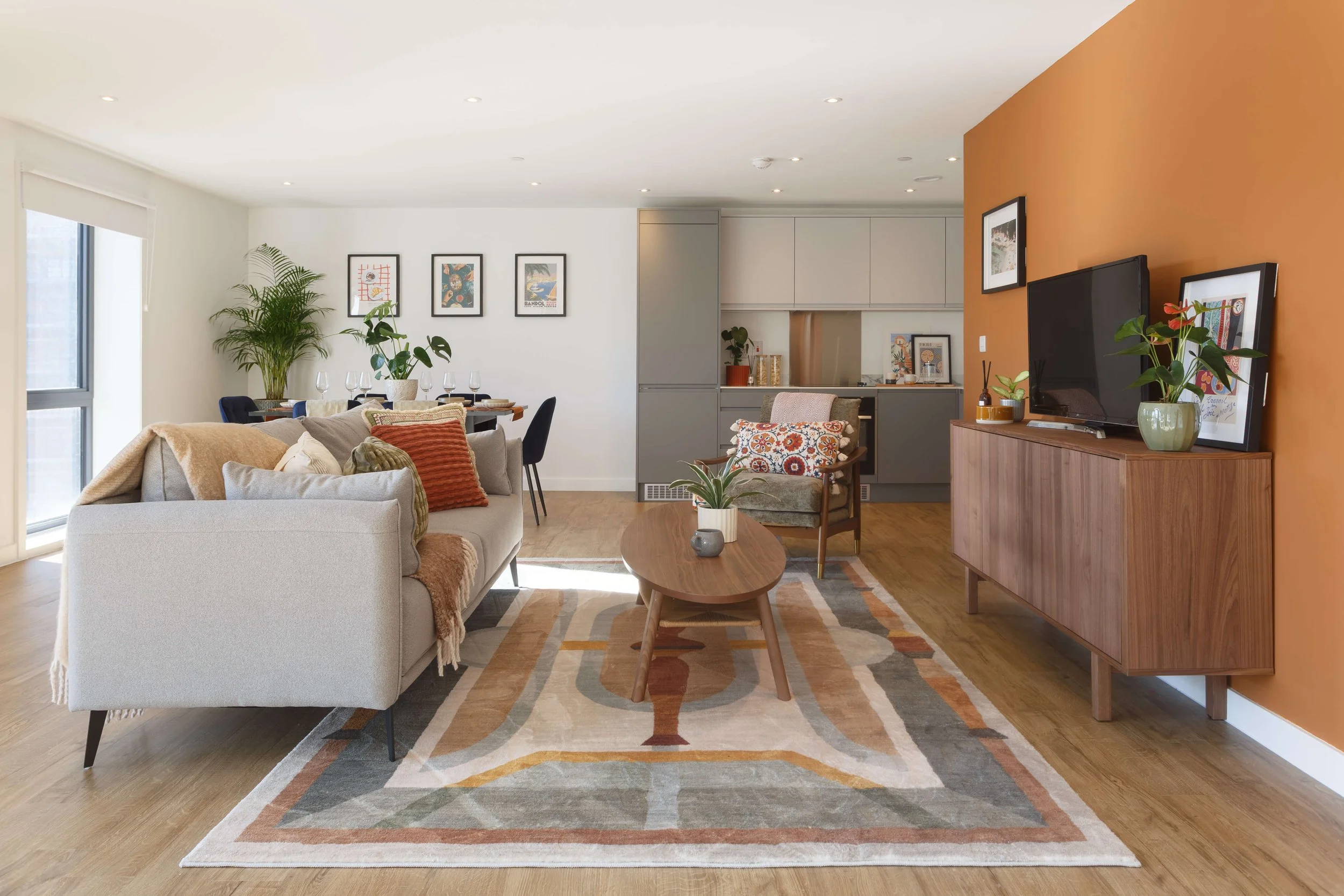 Modern living room with beige sofa, orange accent wall, patterned area rug, wooden coffee table, TV on wooden console, and artwork on the walls.