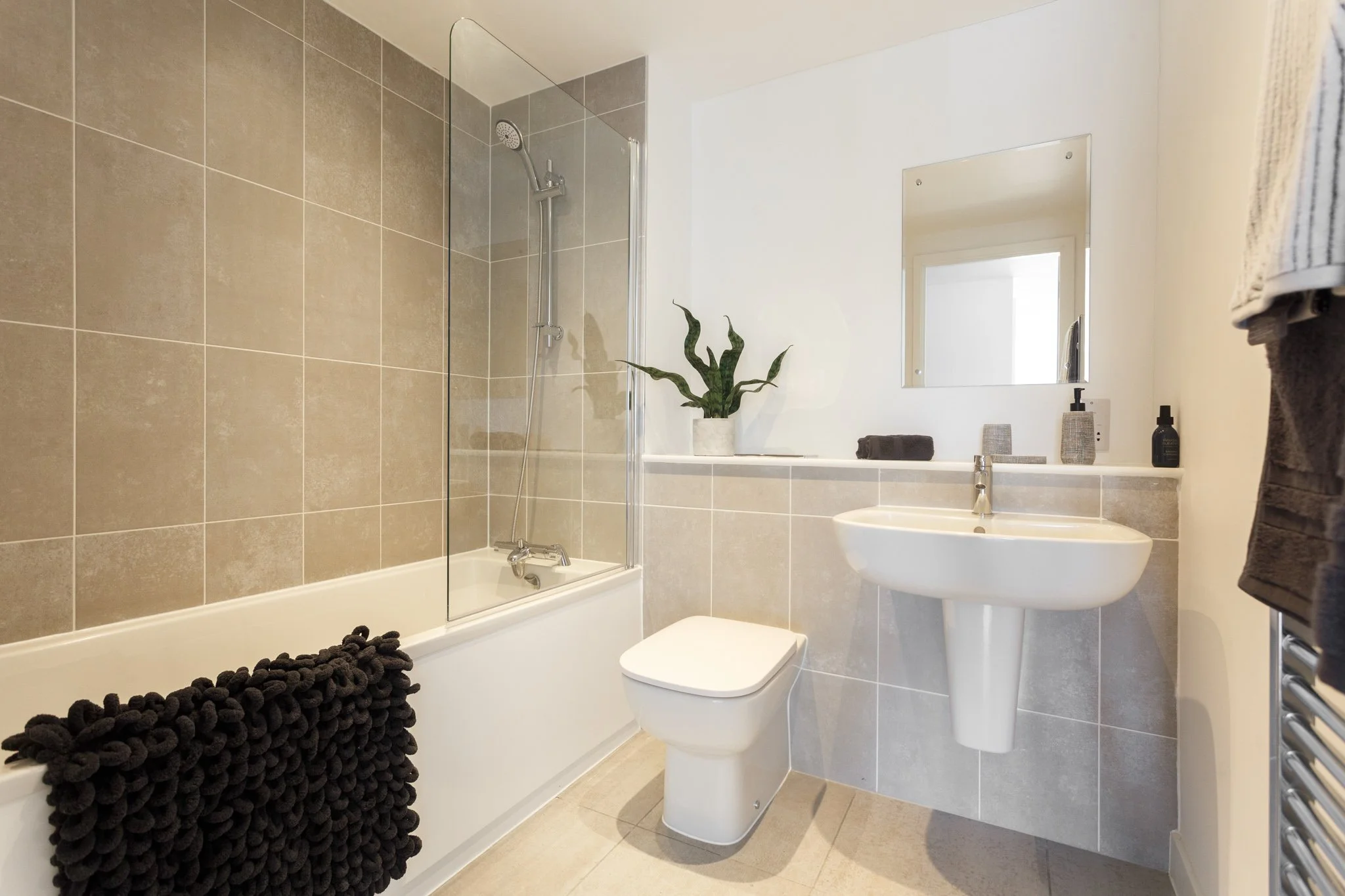 A modern bathroom with beige tiles, a bathtub with a glass shower screen and a handheld showerhead, a white toilet, a white sink, a mirror, a potted plant, two soap dispensers, and a towel hanging on a radiator.