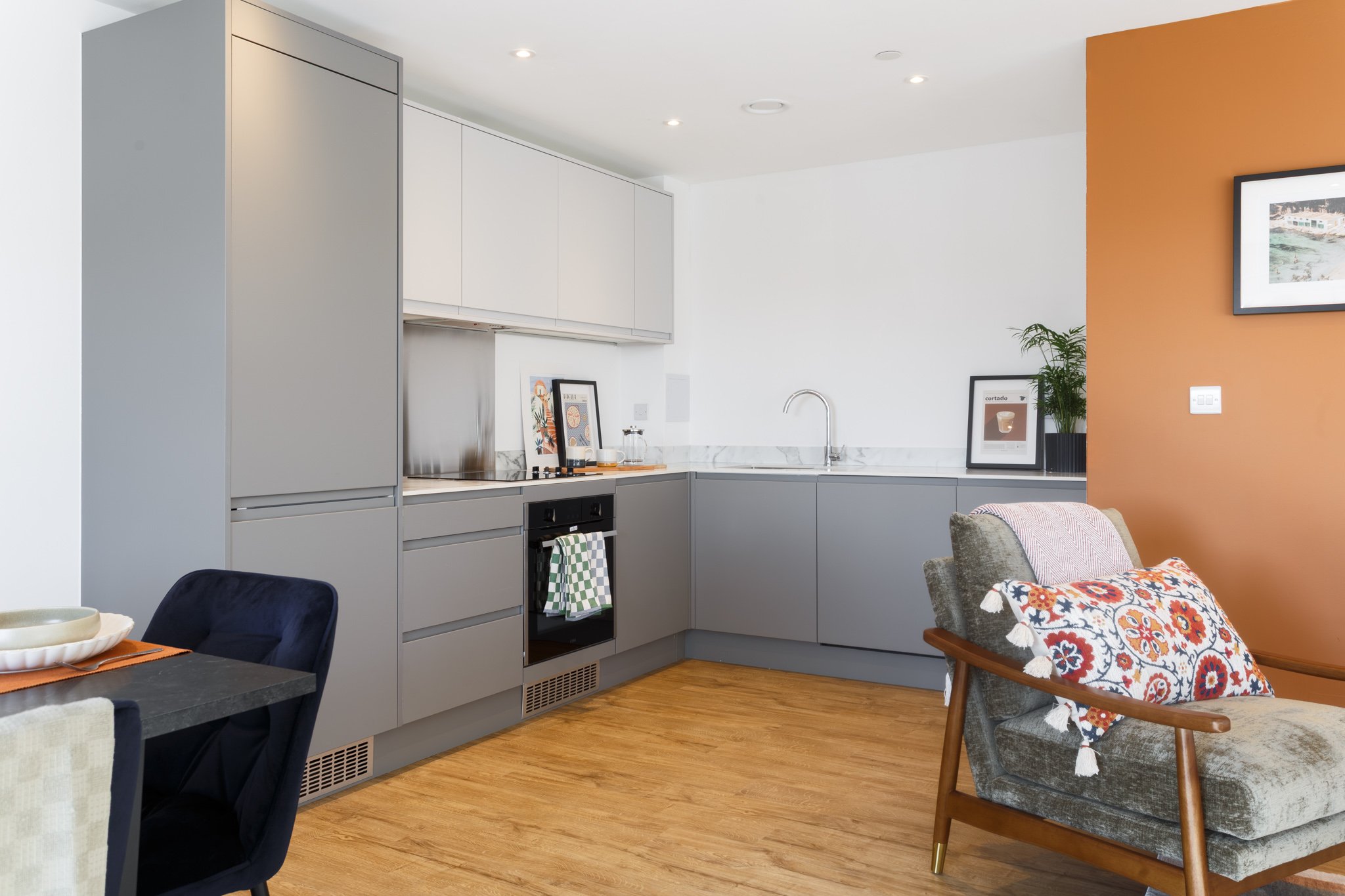 Modern kitchen with white and gray cabinets, a marble countertop, and a stainless steel sink, adjacent to a dining area with patterned chairs and an orange accent wall.