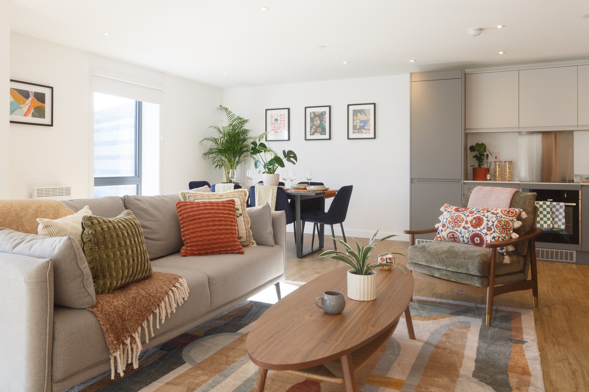 Modern living room with beige sofa, colorful cushions, wooden coffee table, patterned rug, armchair with cushion, dining area with table and black chairs, large window, wall art, plants, and built-in kitchen cabinetry.