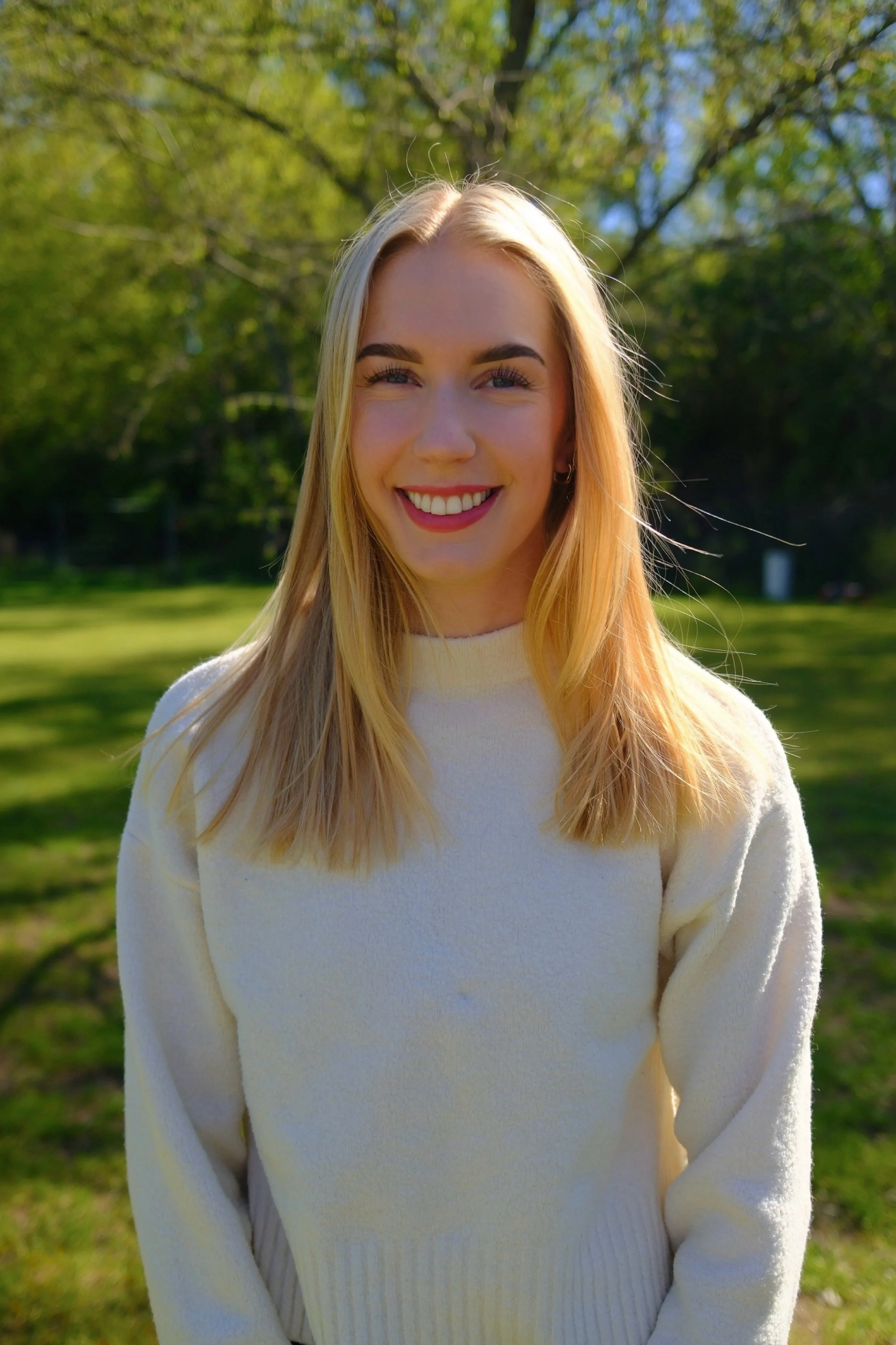 Ein junges Mädchen mit blonden Haaren und einem weißen Pullover steht im Park auf grünem Gras, umgeben von Bäumen im Frühjahr bei sonnigem Wetter.