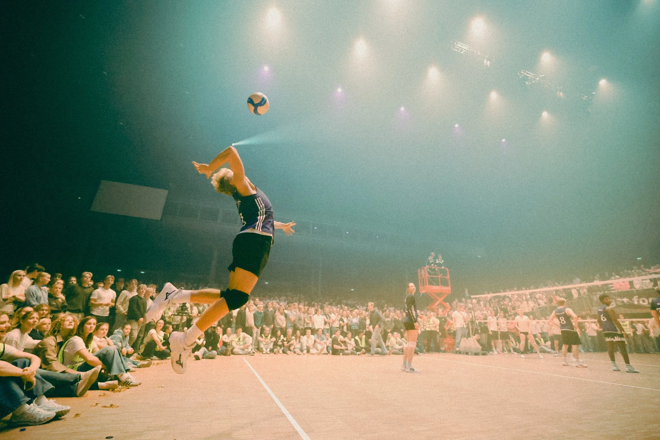 En volleyballspiller hopper for å spike ballen over nettet under en kamp, med en stor tilskuerskare som ser på i en innendørs sportsarena.