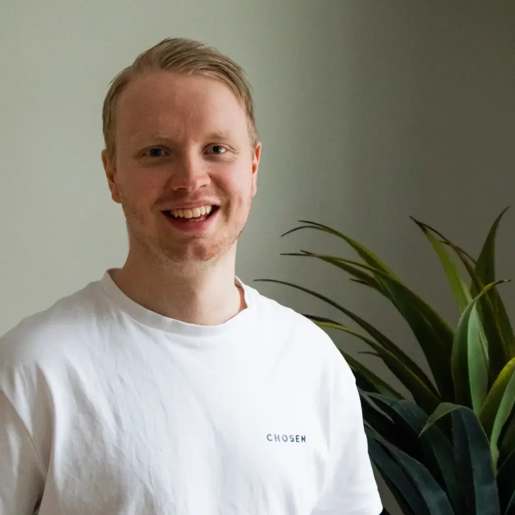 En smilende mann med lyst hår og skjegg iført en hvit t-skjorte som sier "CHOSEN", står foran en grønnsakplante med lange grønne blader på en vegg med svak belysning.