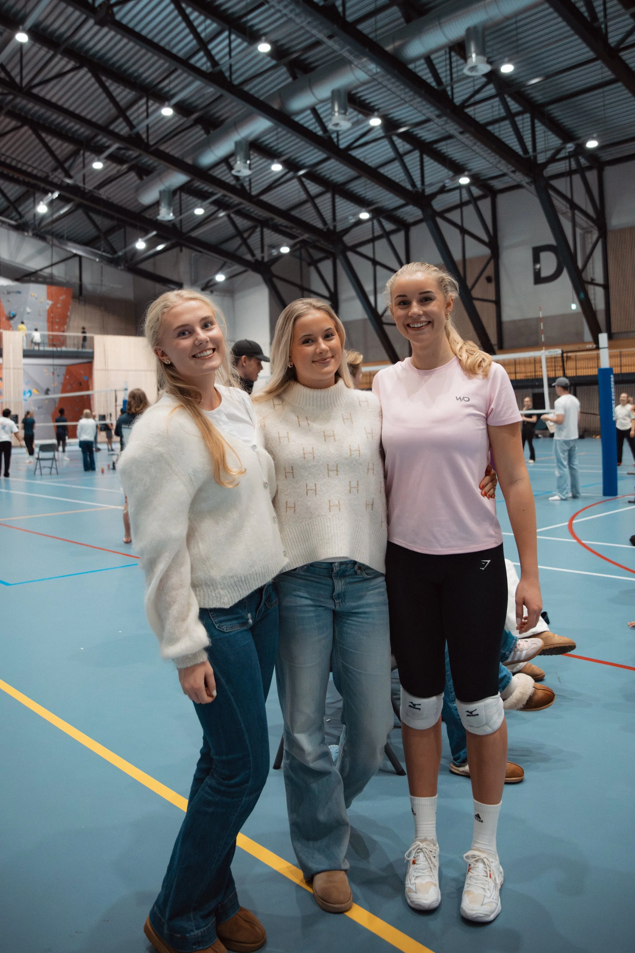 Tre kvinner smiler og står sammen i en innendørs volleyballhall, med volleyballnett og andre personer i bakgrunnen.