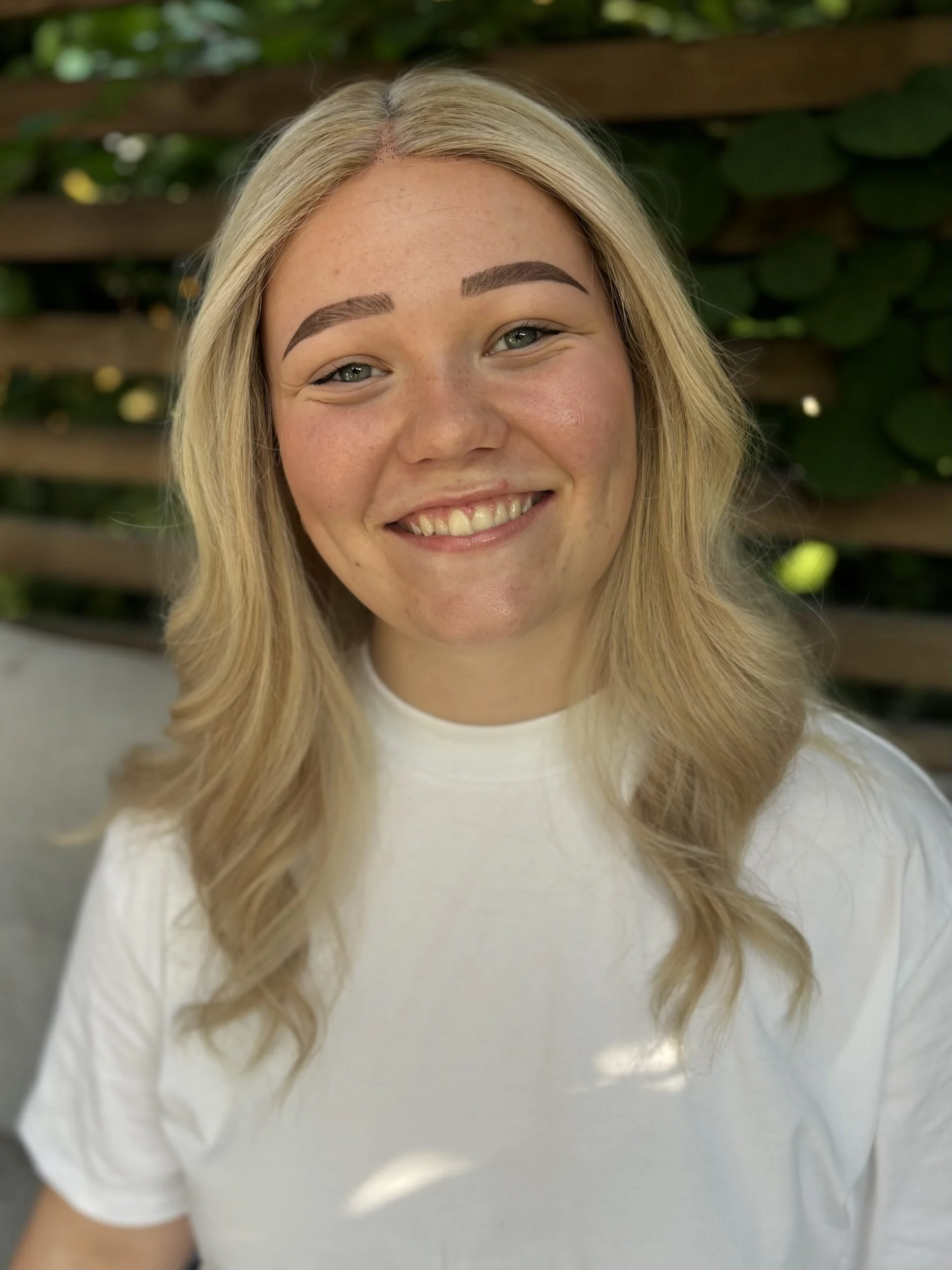 Nærbilde av en kvinne med blondt, bølgete hår, lett smil, lette øyne og naturlige ansiktstrekk, iført hvit genser, med en grønn bakgrunn.