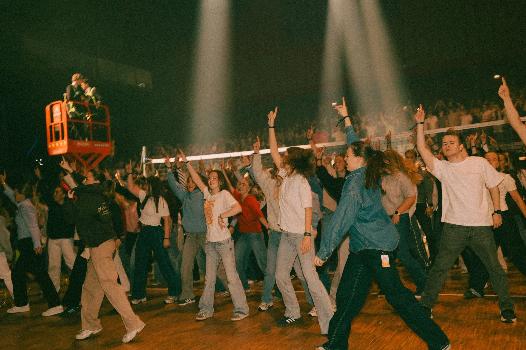 En stor gruppe mennesker danser i en mørk innendørsarena med flere lysstråler som lyser ned på dansegulvet.