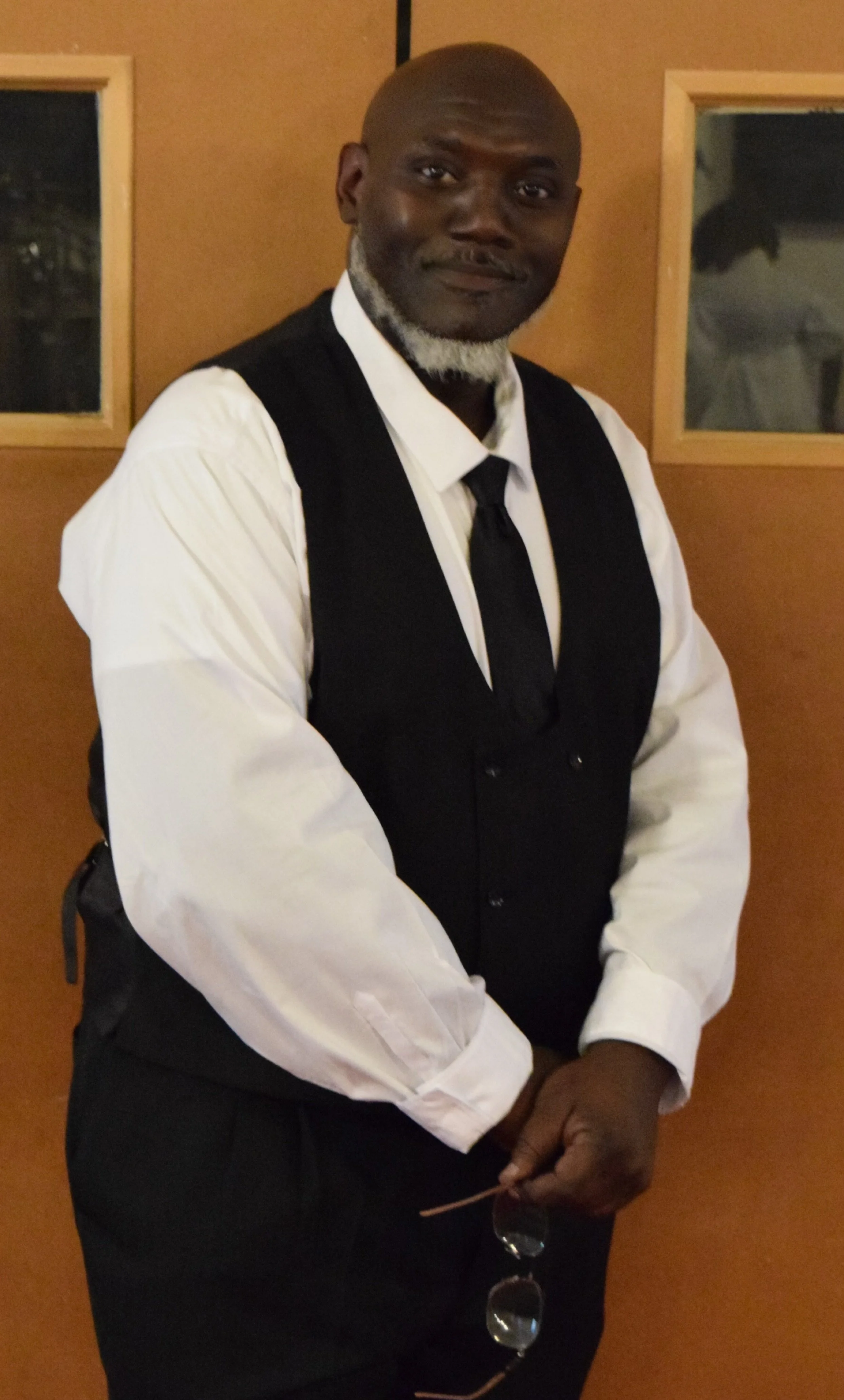 A man dressed in a white shirt, black vest, and tie standing indoors in front of a wooden wall with small windows, holding glasses and a swing.