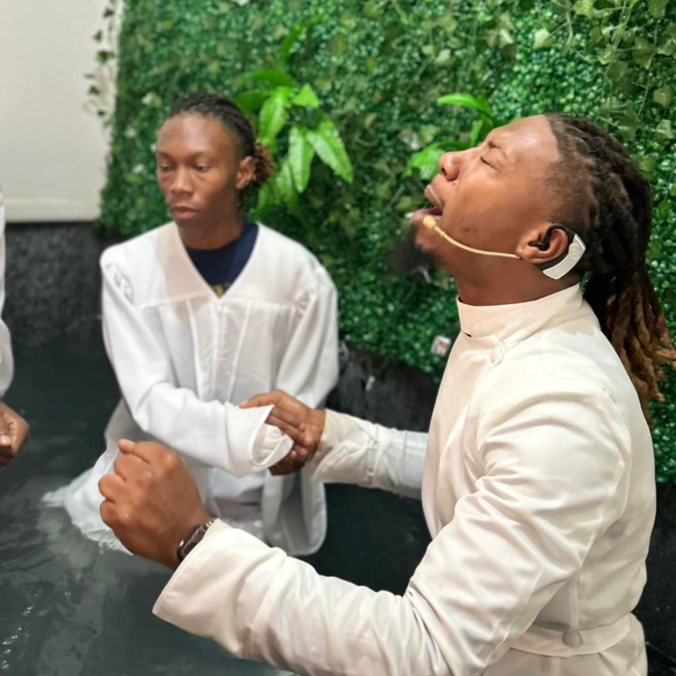 A man with dreadlocks and a headset in a white shirt is baptizing a young woman in a baptismal pool. The woman has braided hair and is wearing a white baptism gown. They are indoors with a wall of green plants in the background.