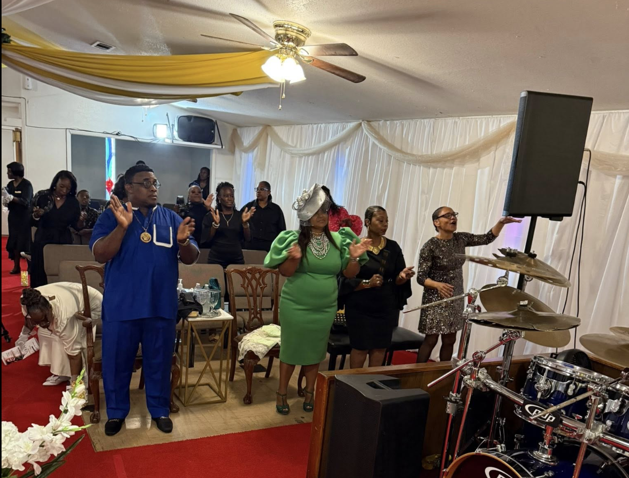 People standing, clapping, and singing in a decorated indoor venue, with a drum set in the foreground. Some individuals are dressed formally, and there are drapes and yellow fabric decorations hanging from the ceiling.